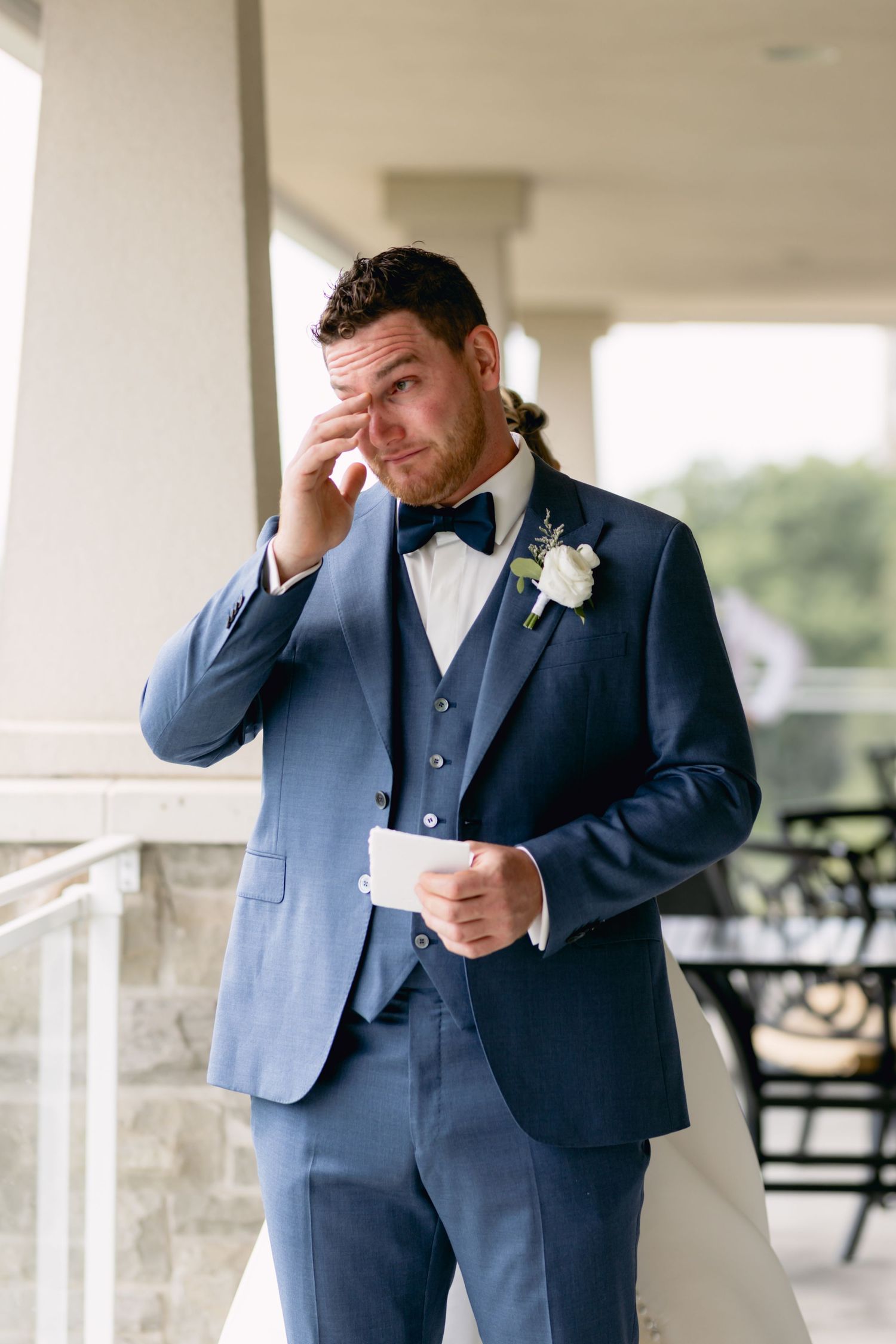 Groom in navy suit becomes emotional while reading note on covered balcony.