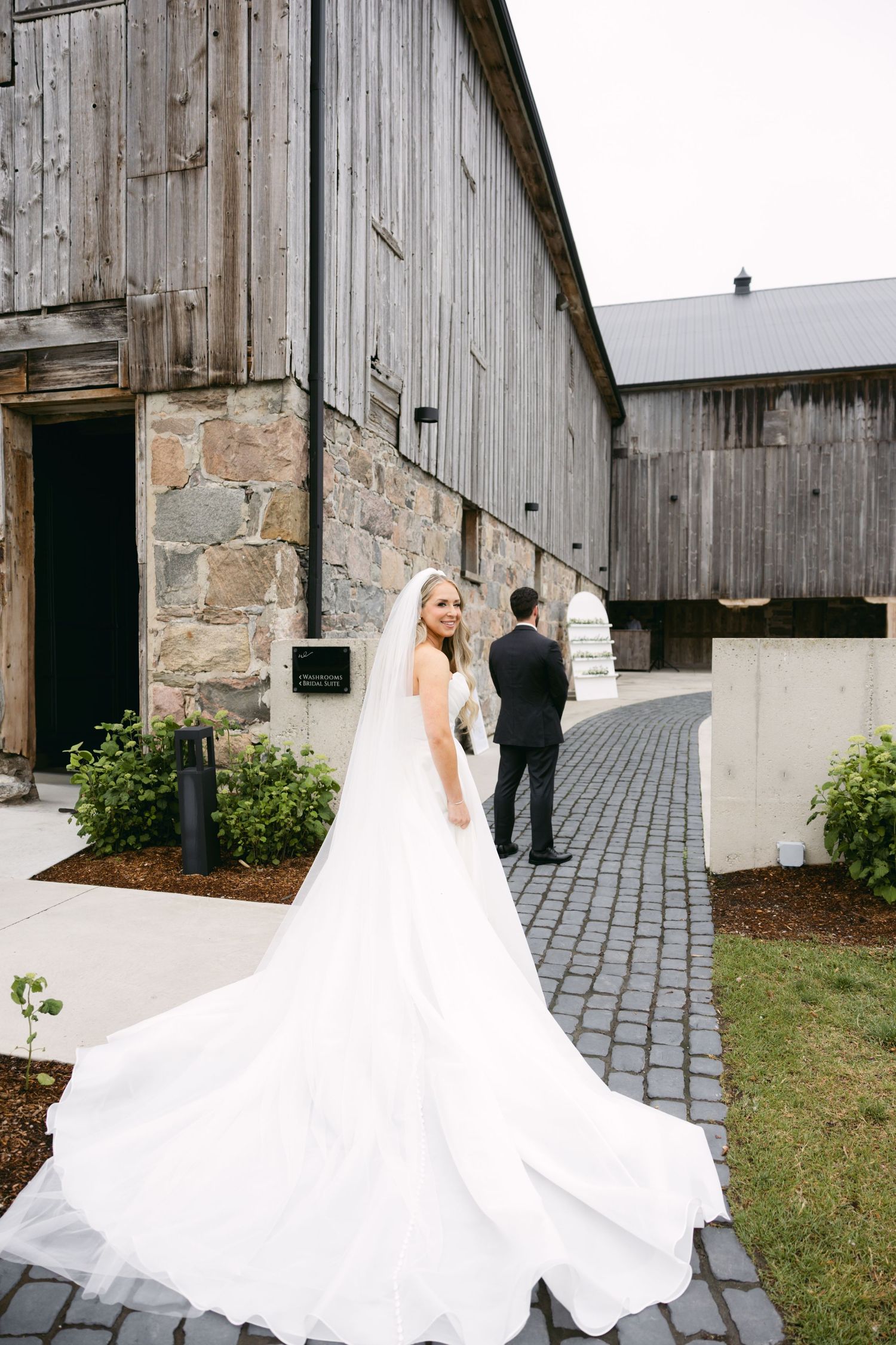 Bride in flowing white gown poses near rustic barn with stone foundation.