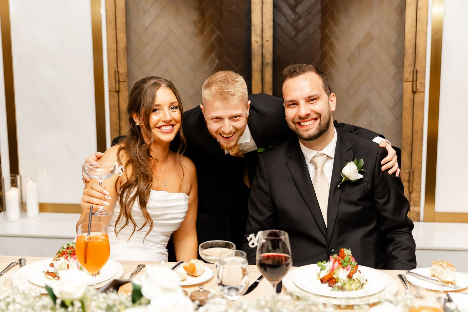 Smiling bride and groom pose with a blond male guest at their elegantly decorated wedding reception table.