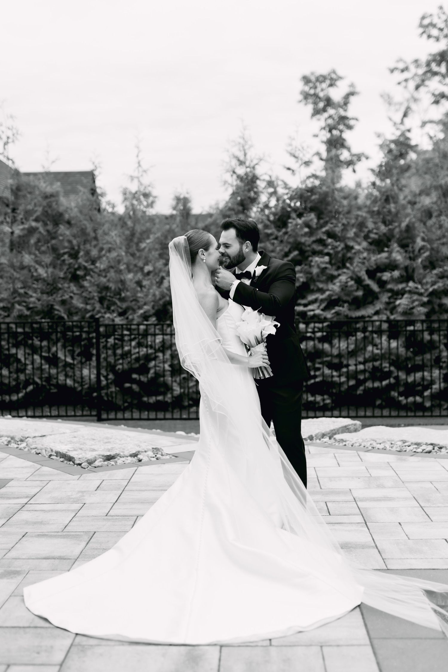 Black and white photo of bride and groom embracing on stone patio with veil flowing behind.