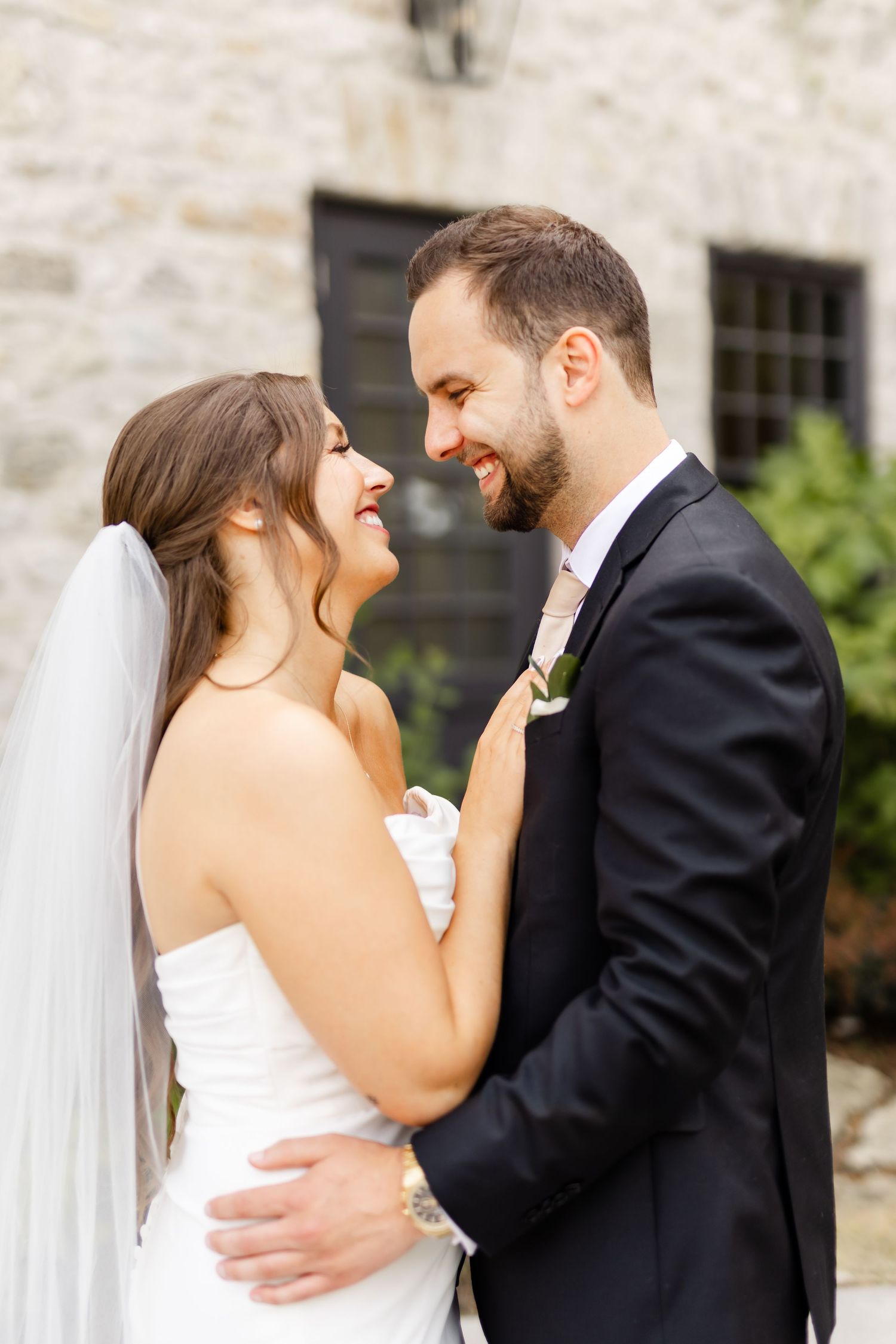Smiling bride and groom embrace closely outdoors, bride in strapless white gown with veil, groom in black suit.