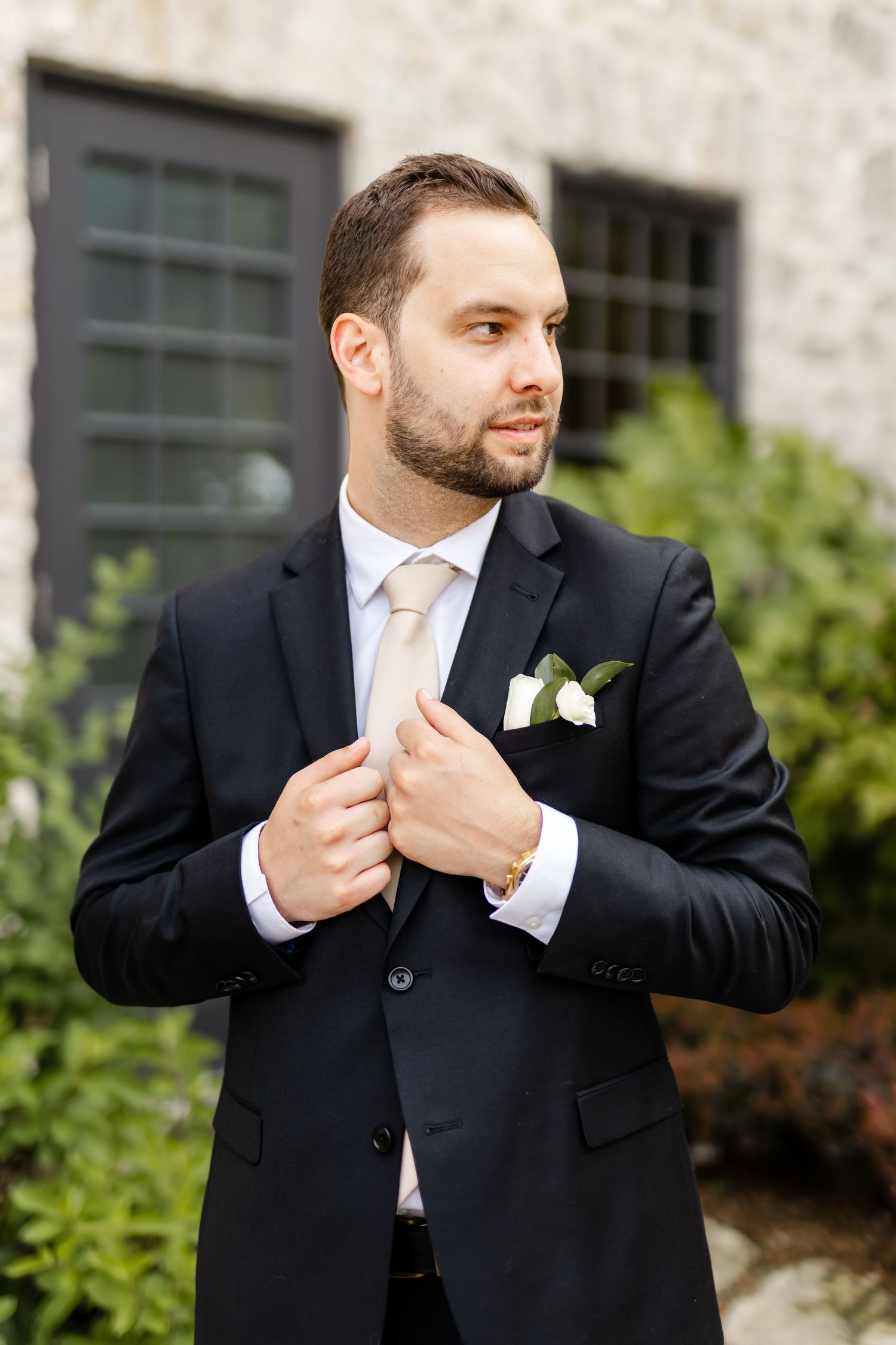 Dapper groom in black suit with white boutonniere adjusts his lapel while standing outdoors near greenery.