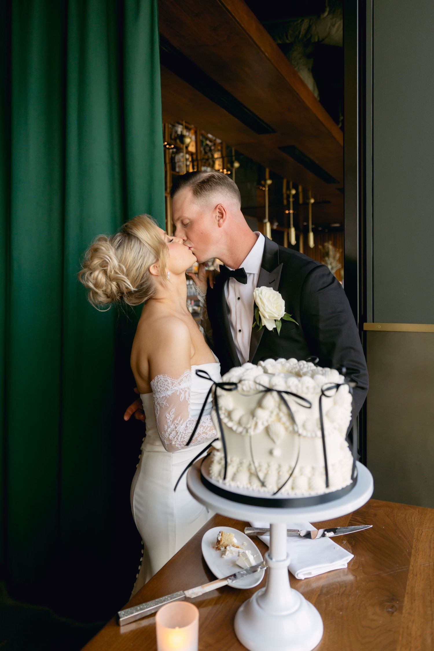 Bride and groom sharing a kiss behind white wedding cake with black ribbon decorations.
