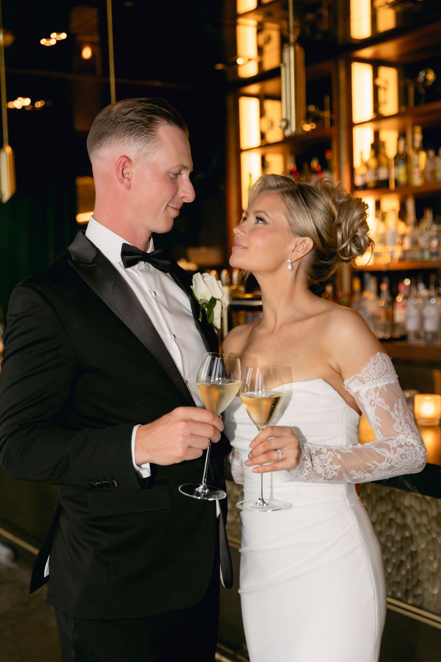 Bride and groom in formal attire toast with champagne glasses at a warmly lit bar.