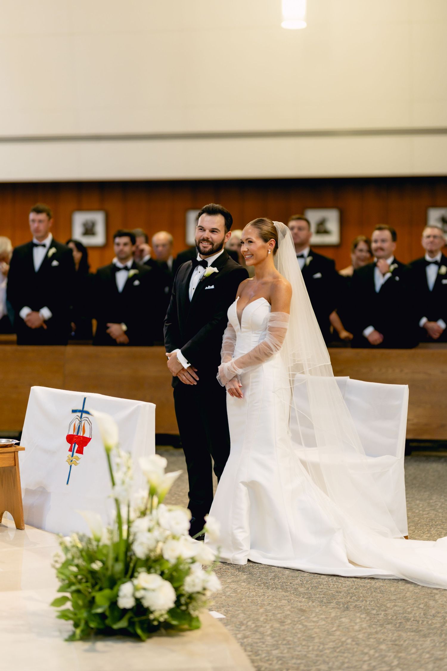 Bride and groom standing at church altar smiling, groomsmen in black tuxedos visible in background.