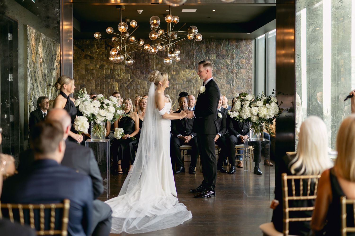 Bride and groom exchange vows during indoor wedding ceremony with gold chandeliers and floral arrangements.