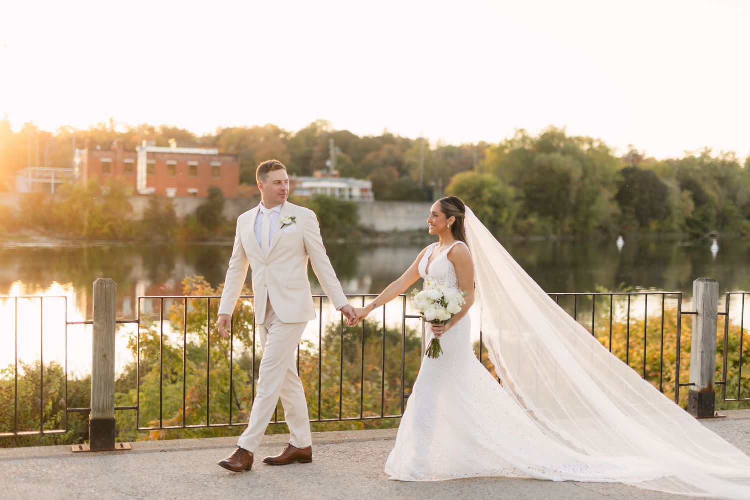 Wedding couple walks along waterfront path during golden hour with veil trailing behind.