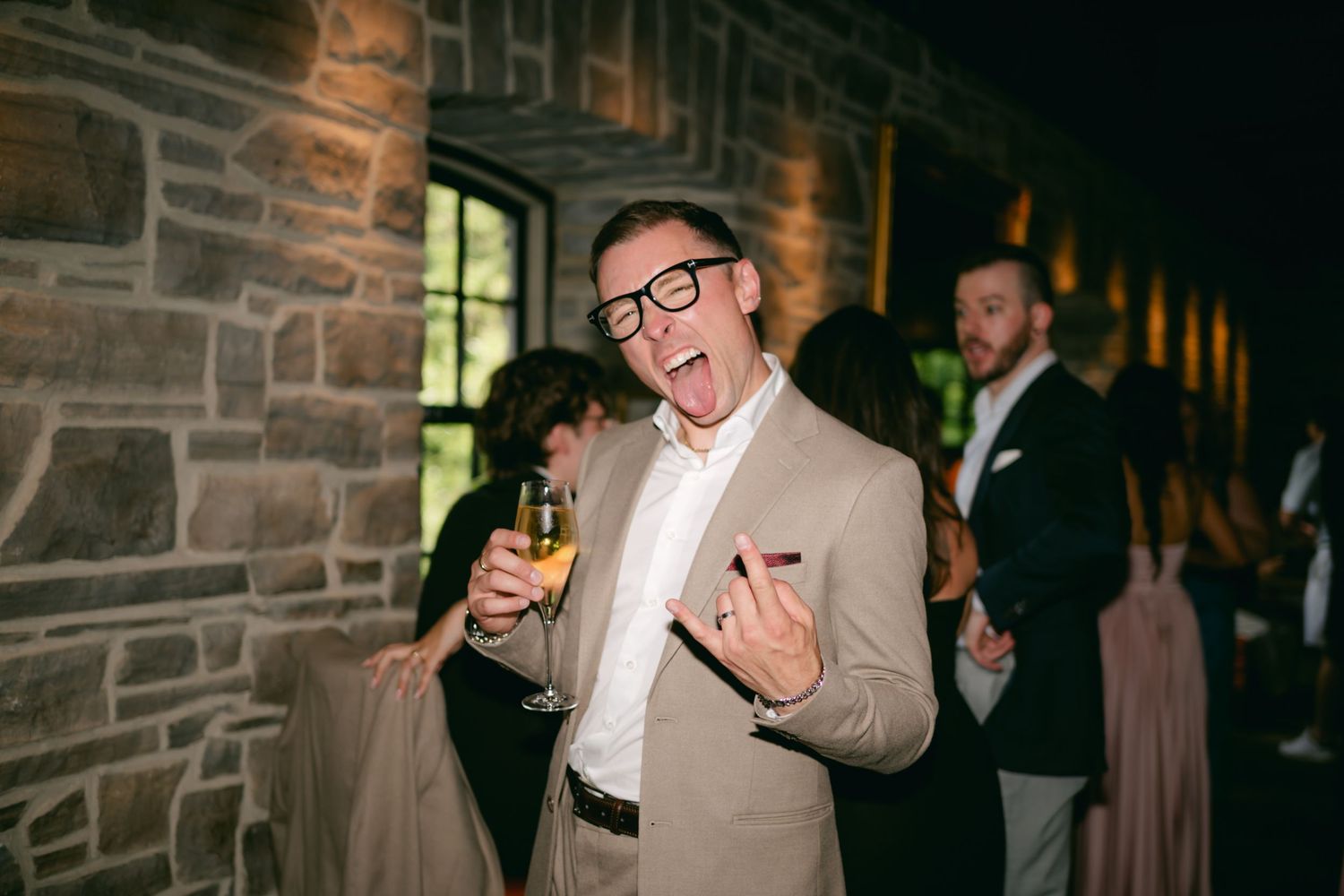 Wedding guest in tan suit celebrating with drinks at stone-walled reception venue.