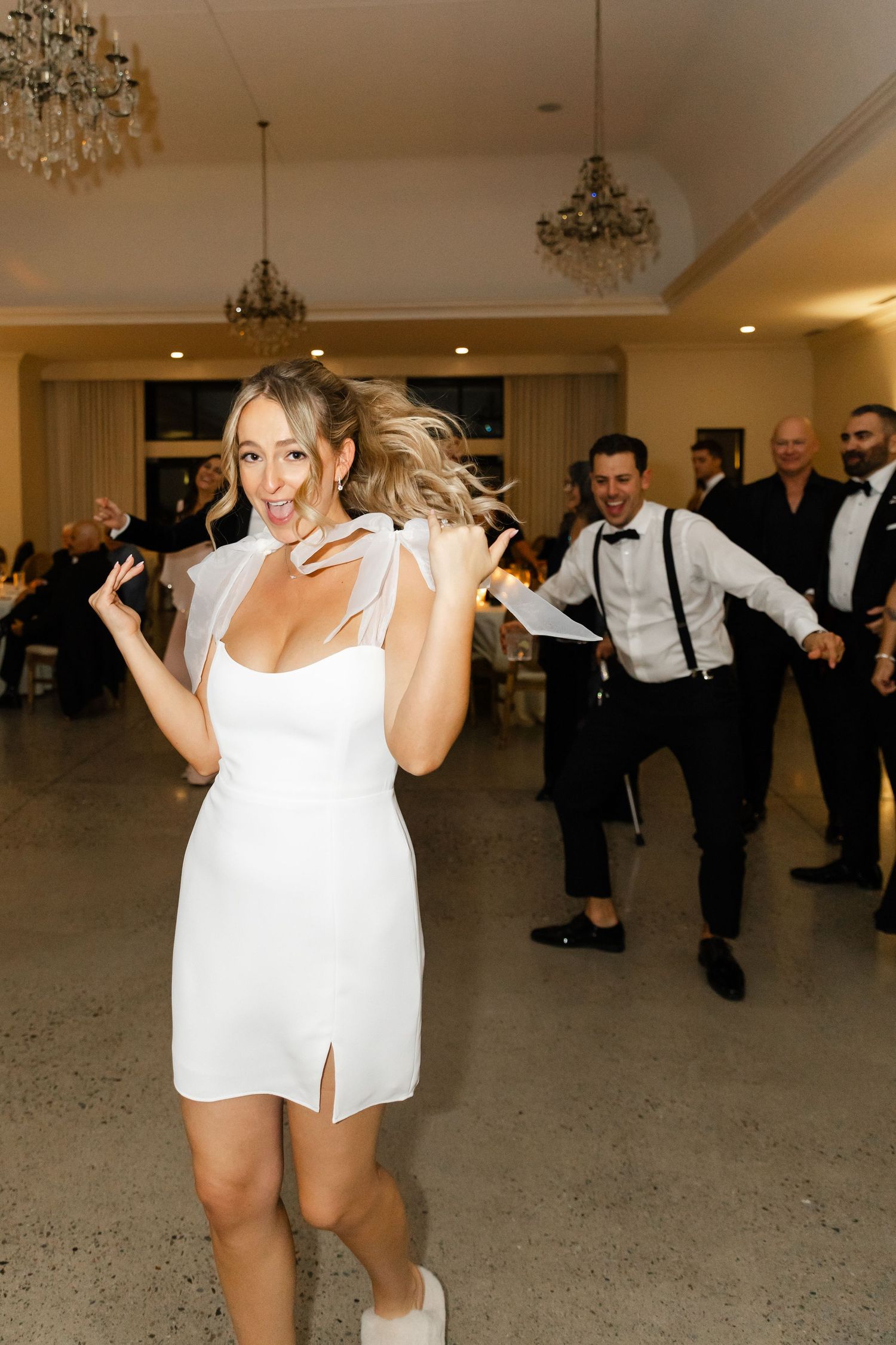 Wedding party dances energetically in white attire on a ballroom floor.