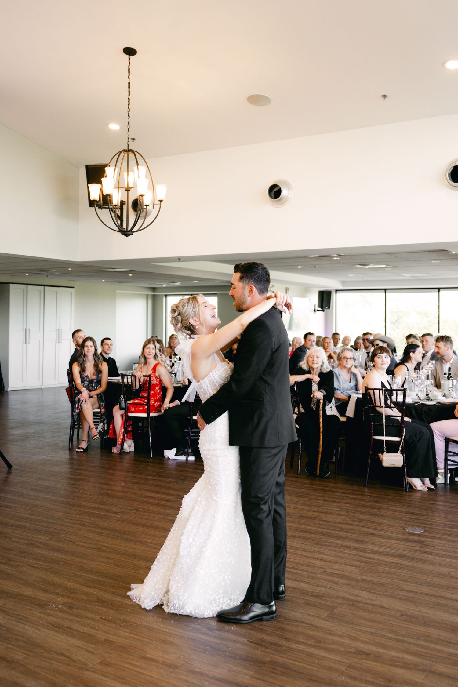 Wedding couple dances in spacious reception hall with hardwood floors and watching guests.