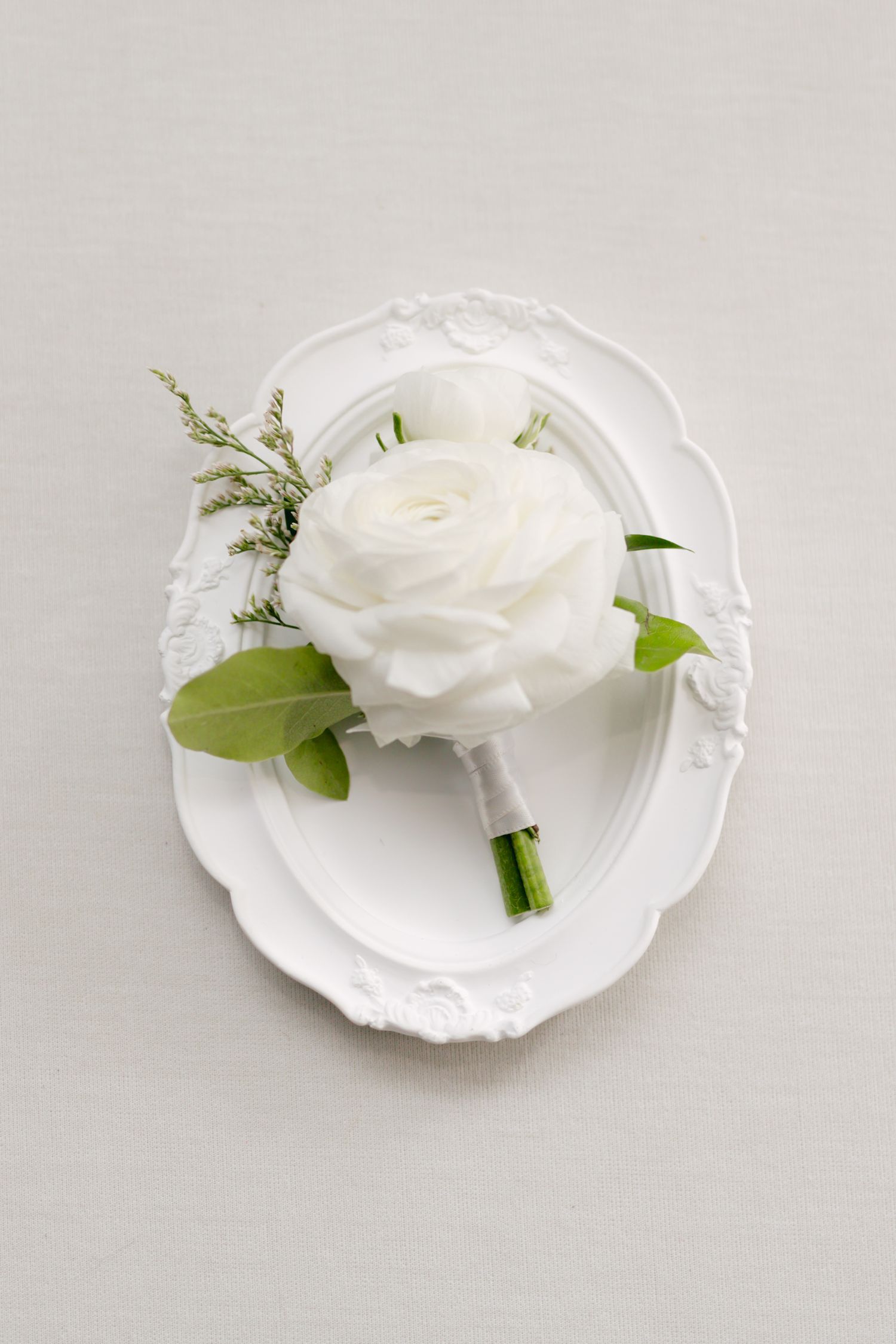 A single white rose boutonniere with greenery displayed on an ornate white plate.