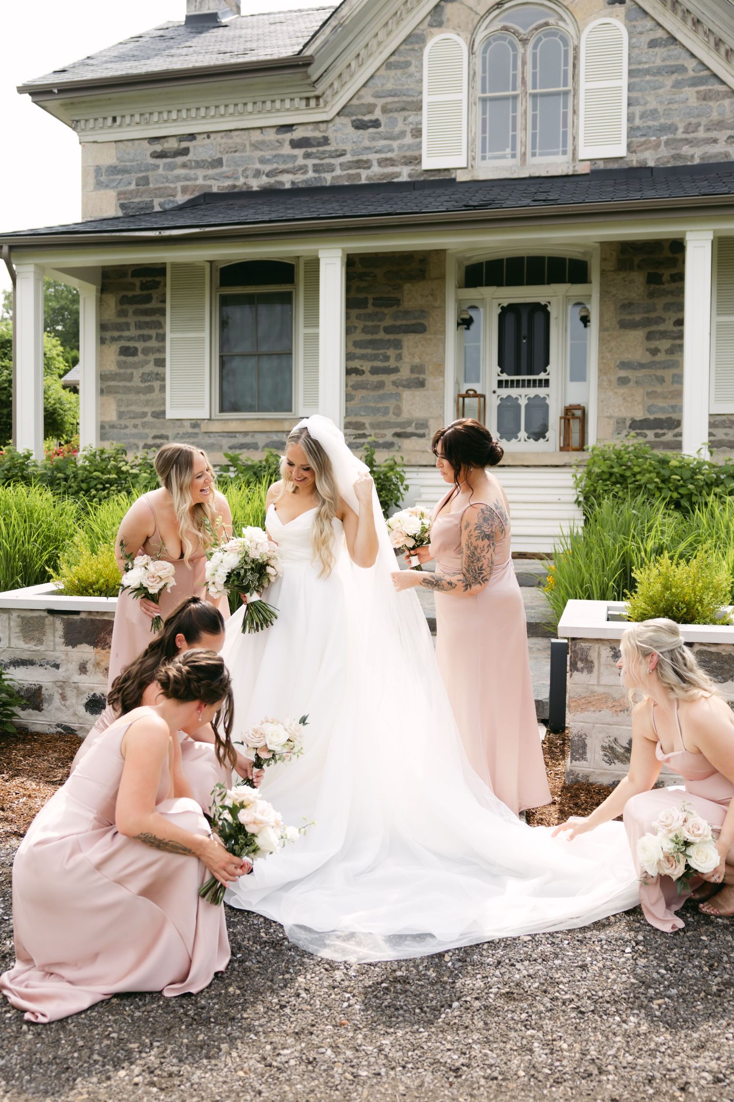 Bridal party in pink dresses sitting on gravel while bride stands in flowing white gown.