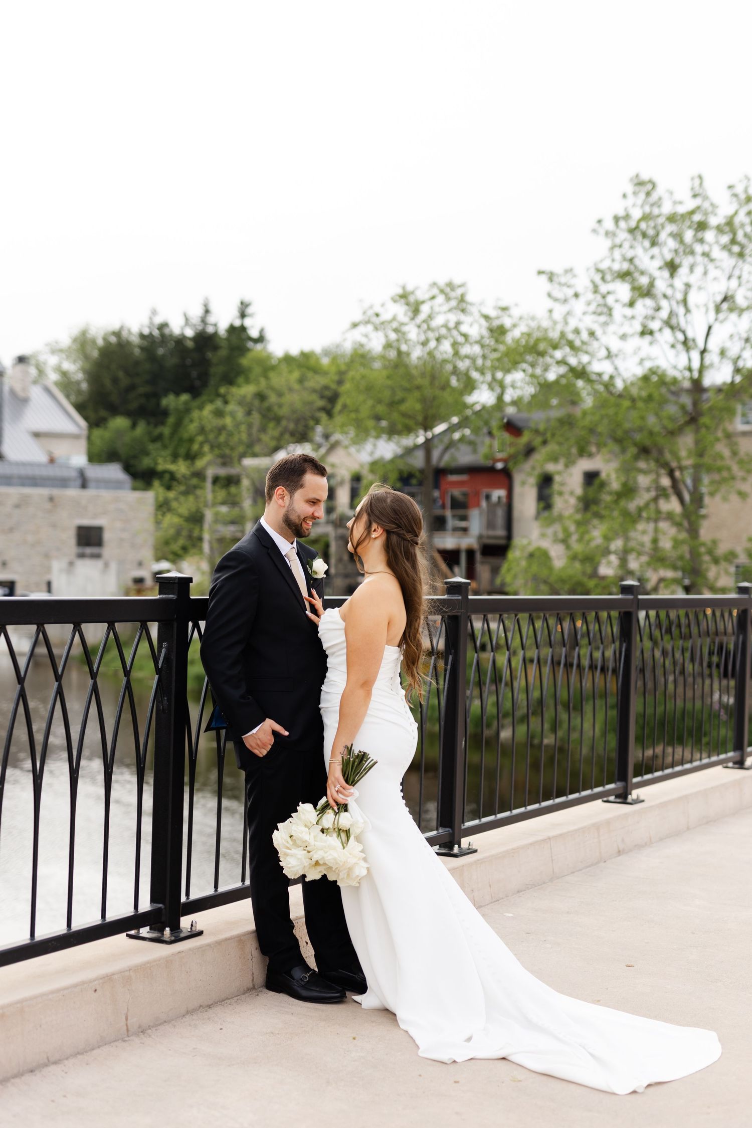 Bride and groom stand romantically on a bridge overlooking a river, bride holding white bouquet.