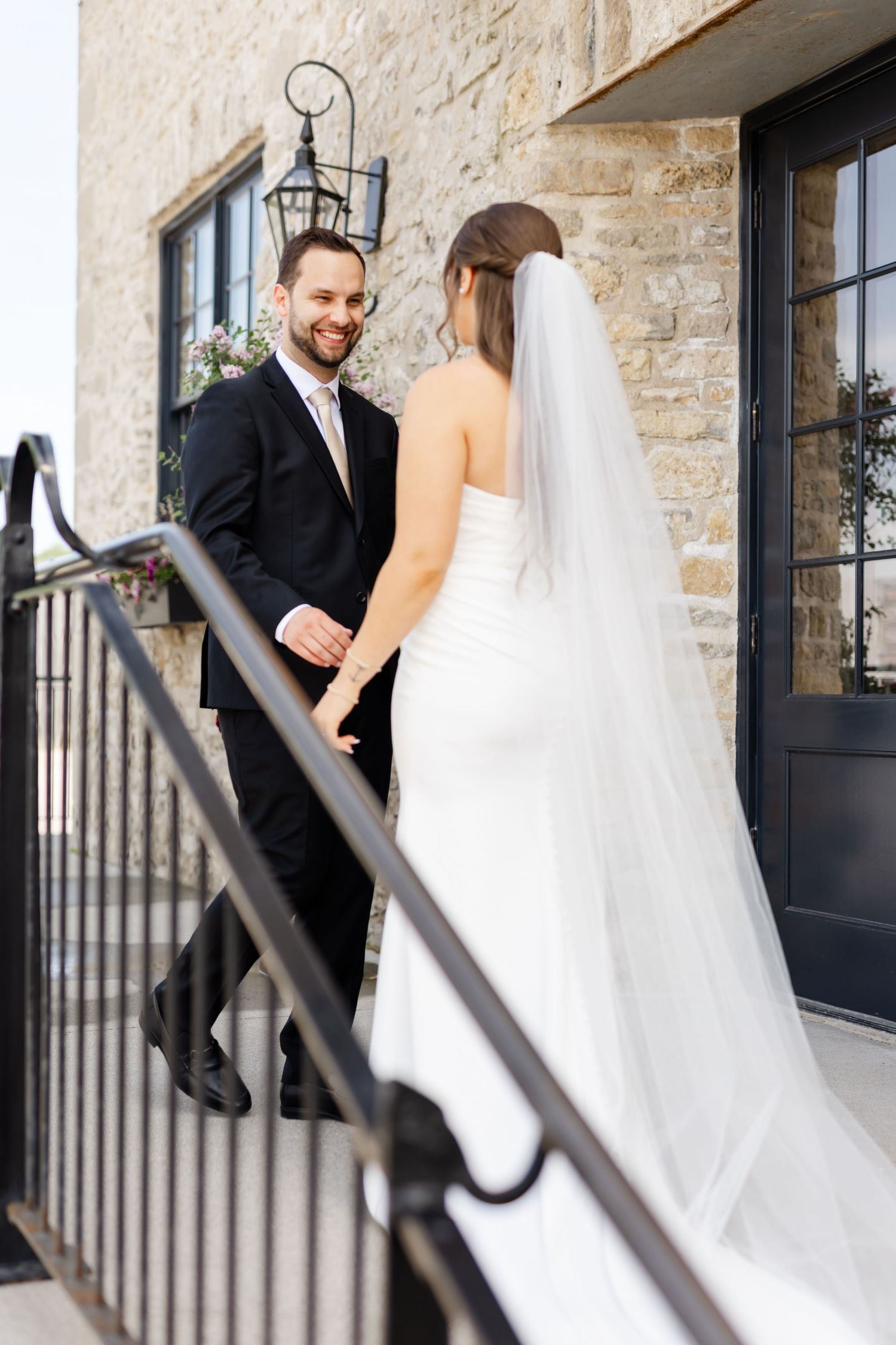 Bride and groom share a joyful moment on stone building steps, groom in black suit, bride in white strapless gown with veil.