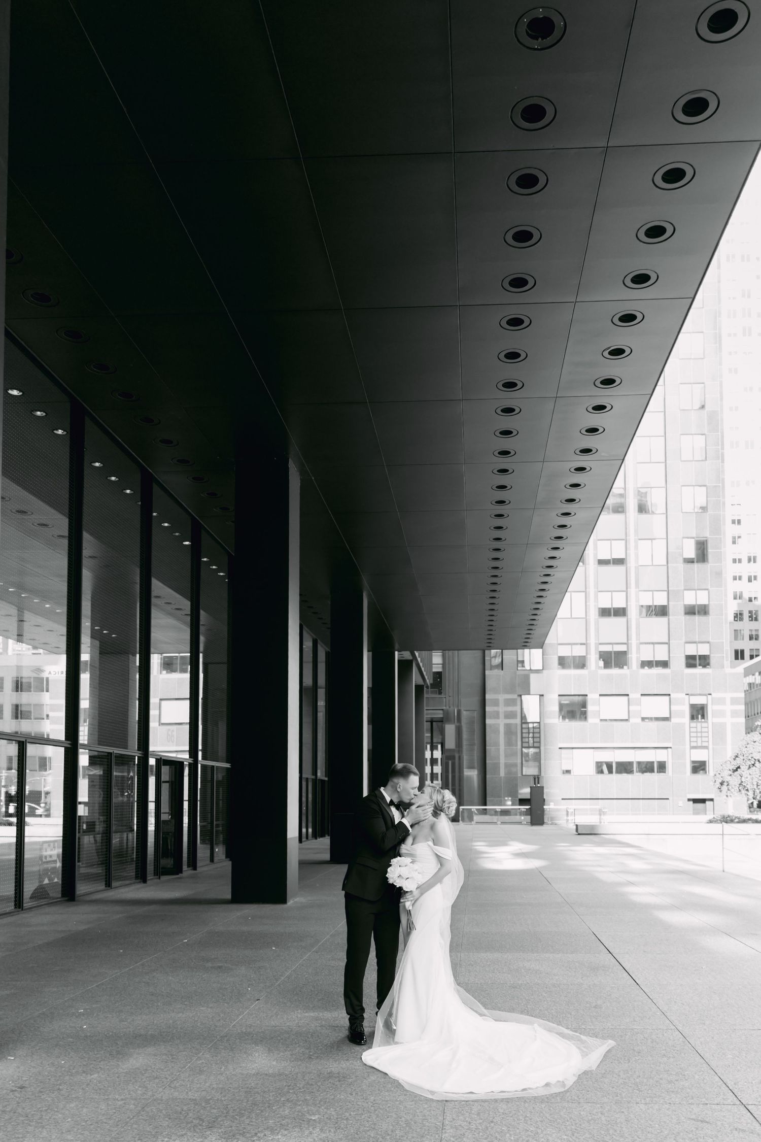 Black and white photo of bride and groom embracing under modern architectural overhang in the city.