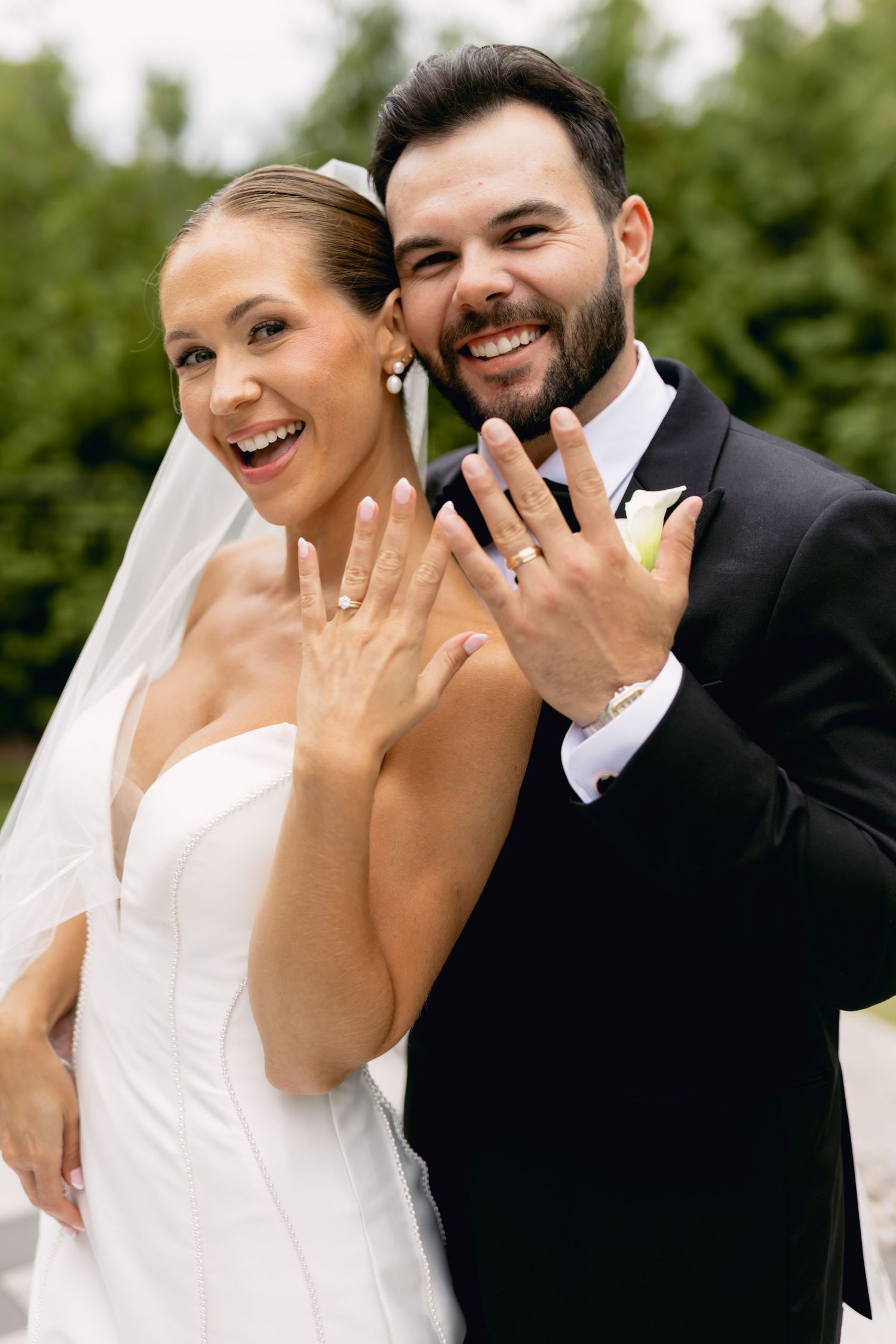 Smiling bride and groom show off wedding rings to camera in outdoor portrait.