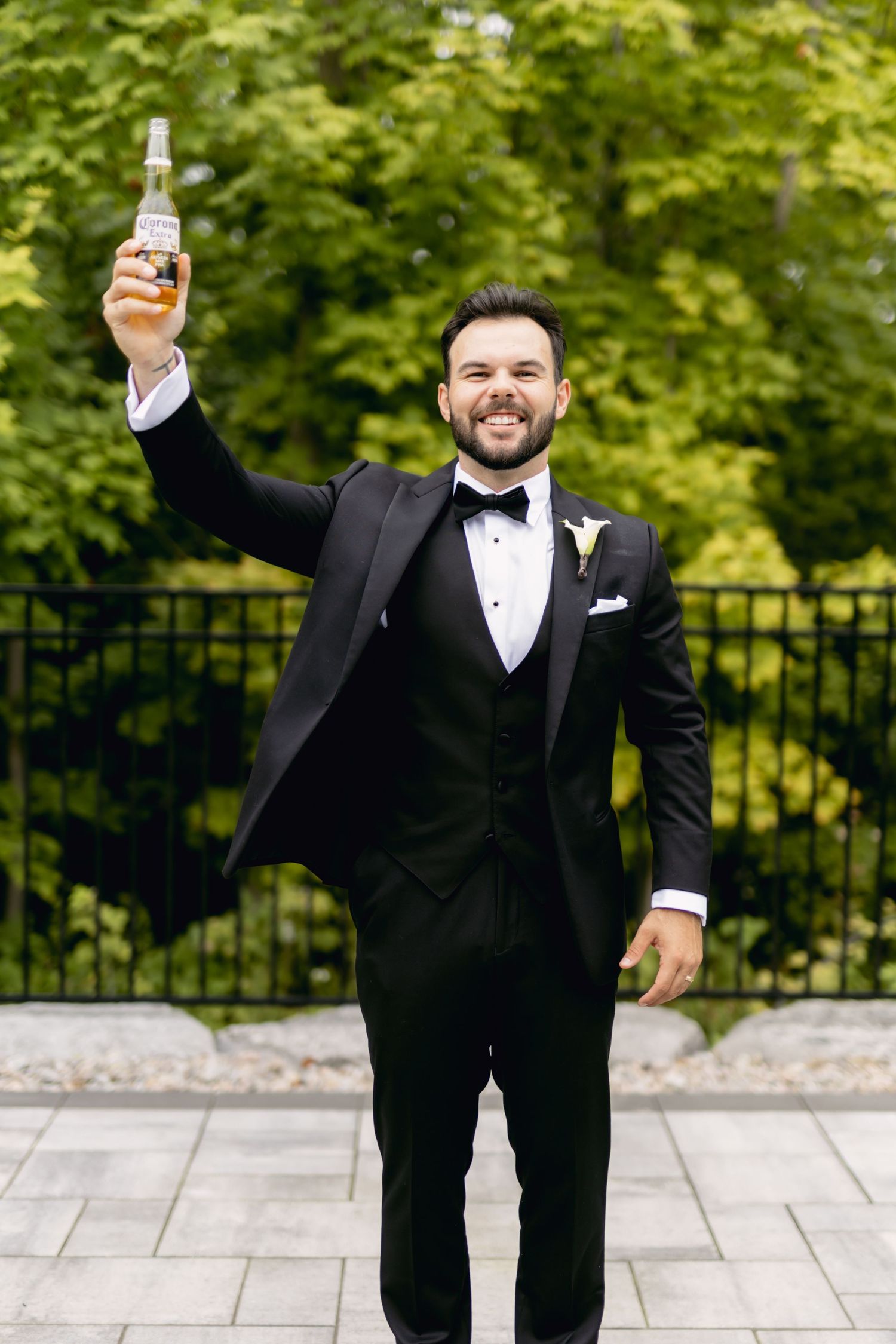 Smiling groom in black tuxedo raises a beer bottle outdoors with green trees behind him.