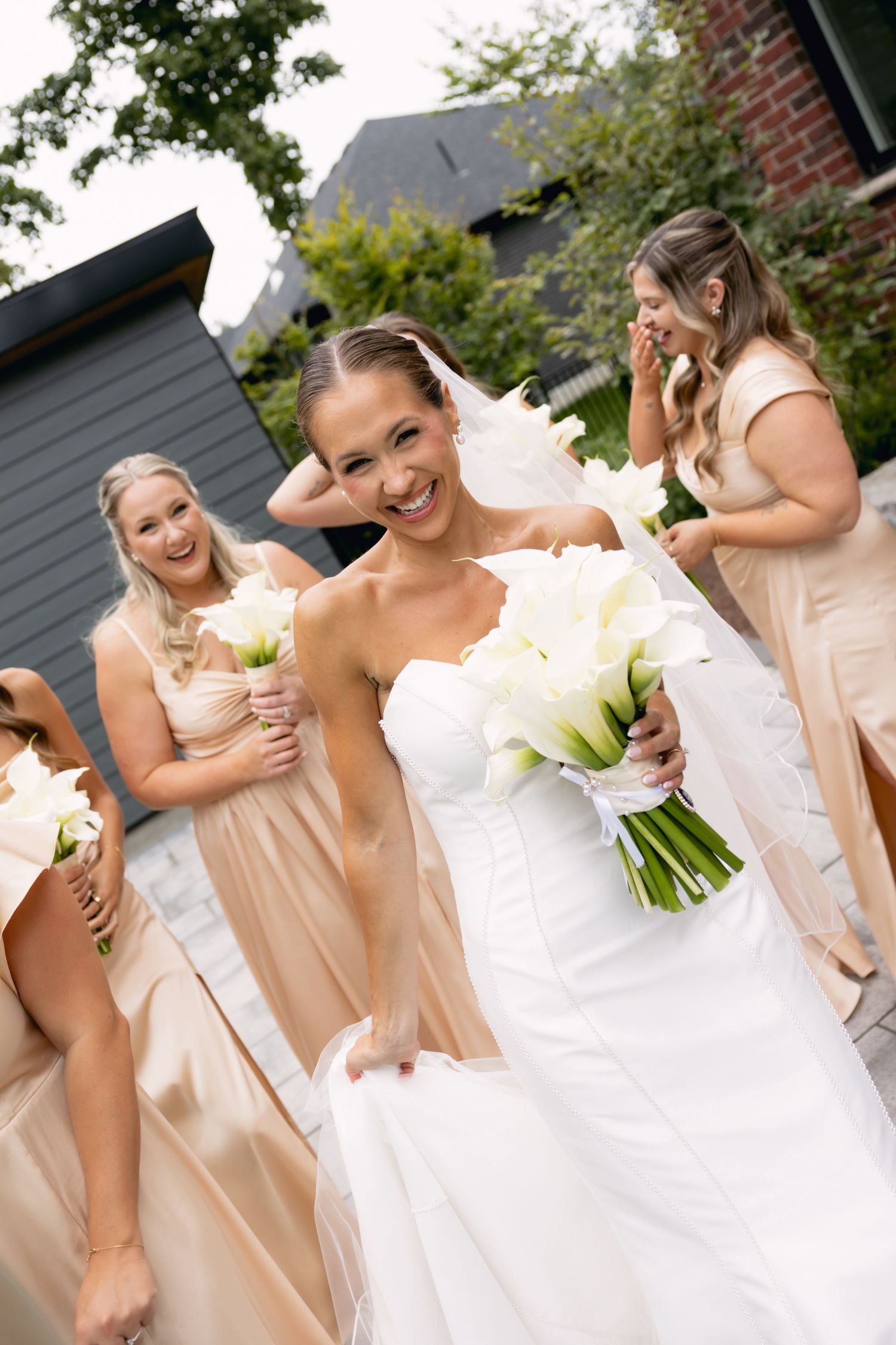 Smiling bride in strapless gown walks with bridesmaids in champagne dresses holding calla lilies.