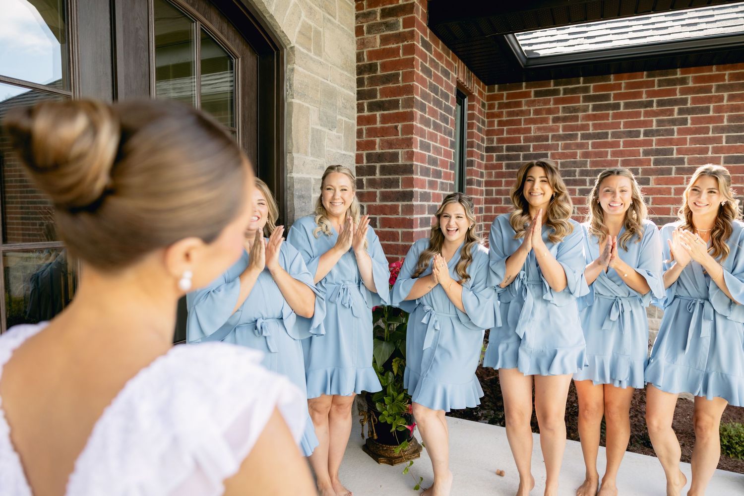Excited bridesmaids in blue robes react to bride's first look on brick building patio.