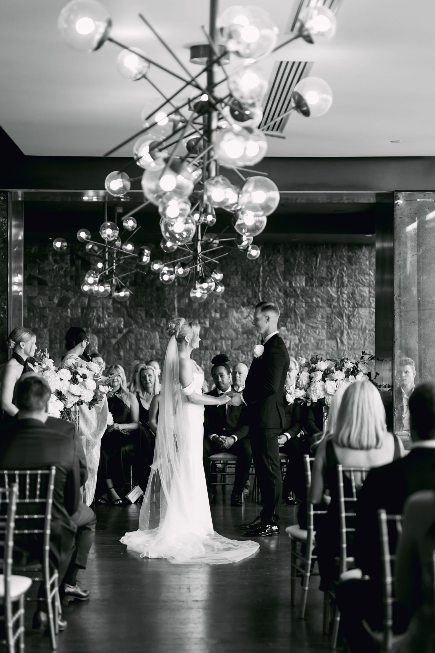 Black and white photo of bride and groom exchanging vows under modern chandelier with seated wedding guests.