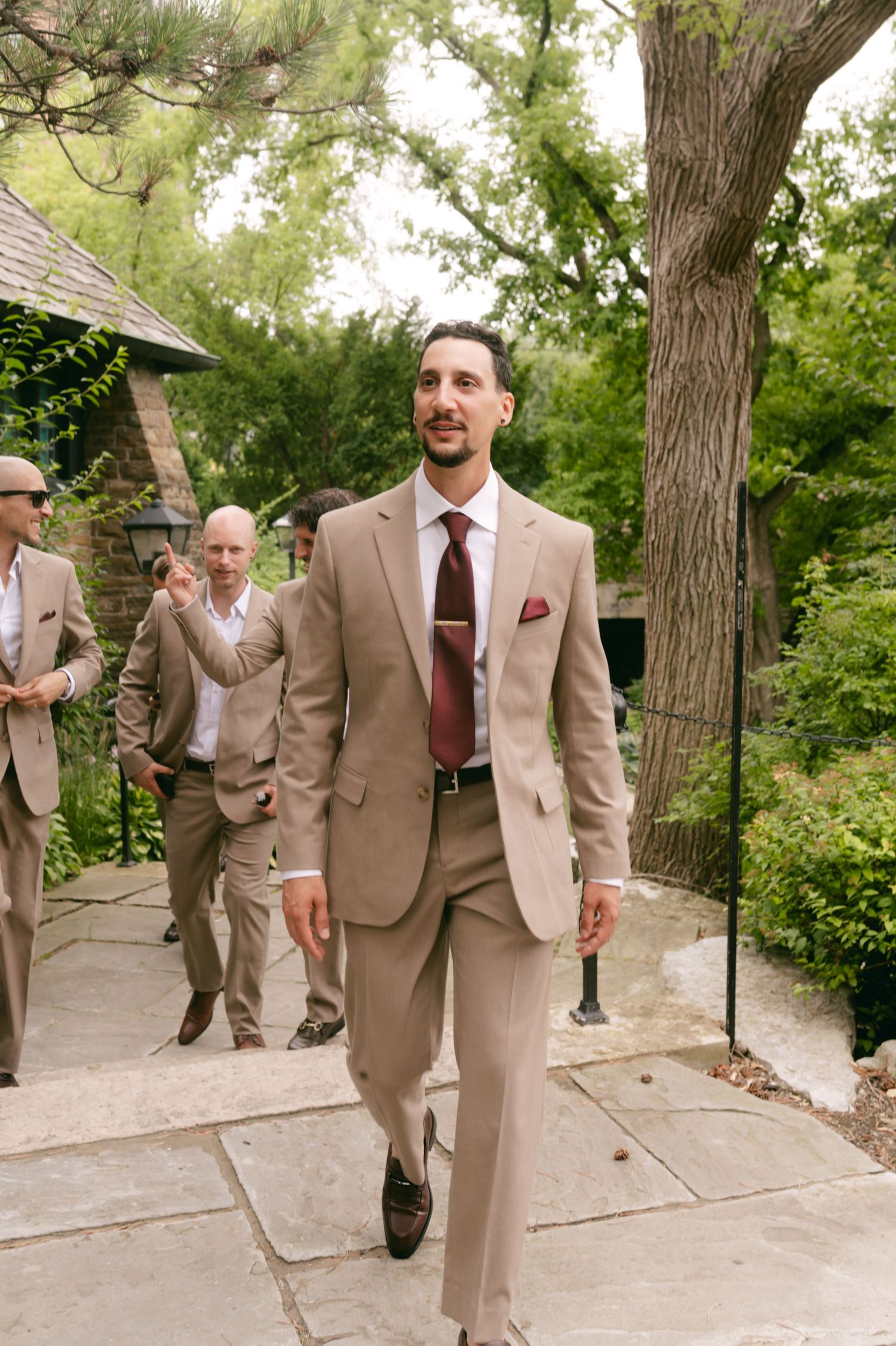 Groomsmen in beige suits walking down an outdoor path surrounded by trees.