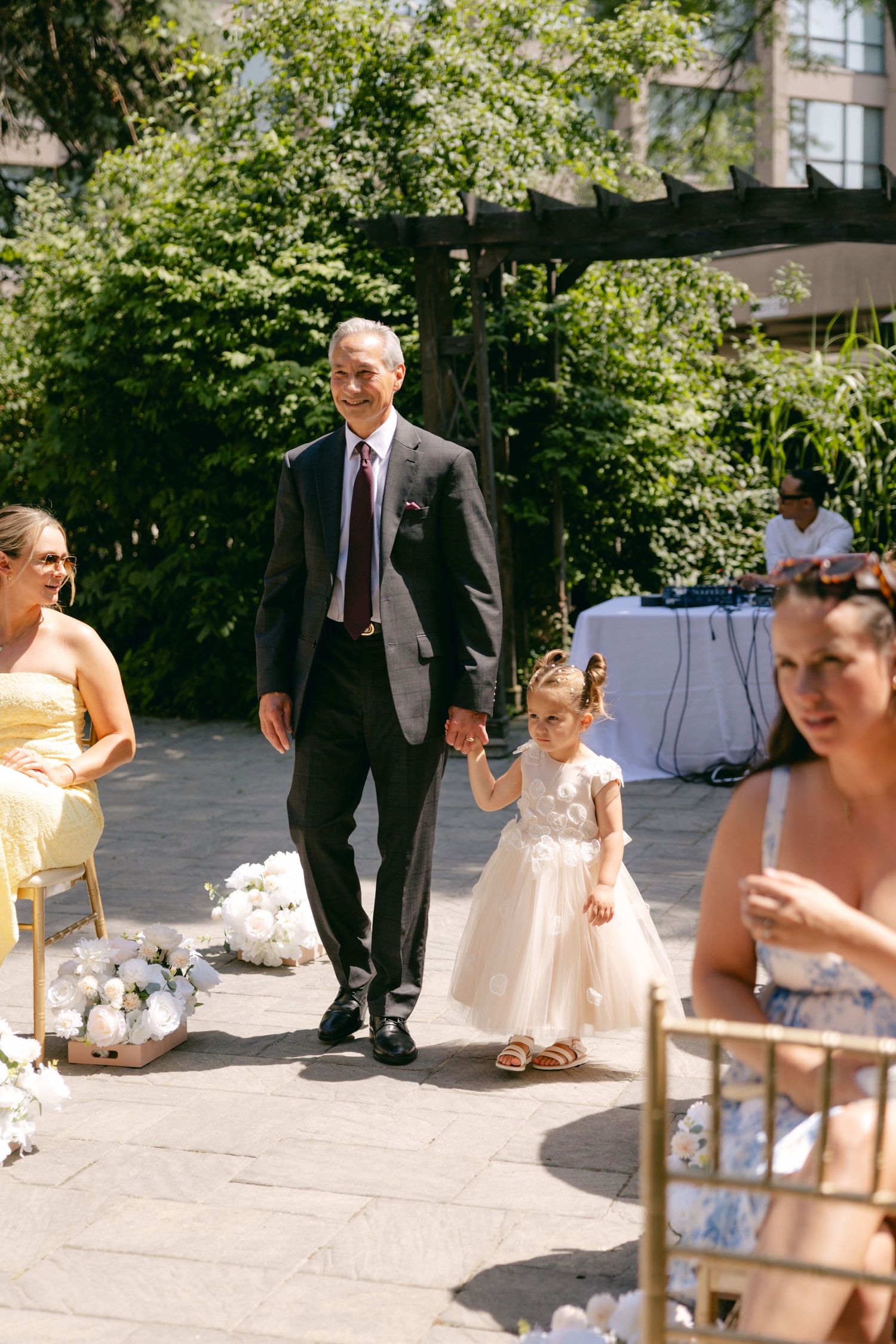 Wedding party members gather outdoors on a sunny day with chairs and decorations visible.