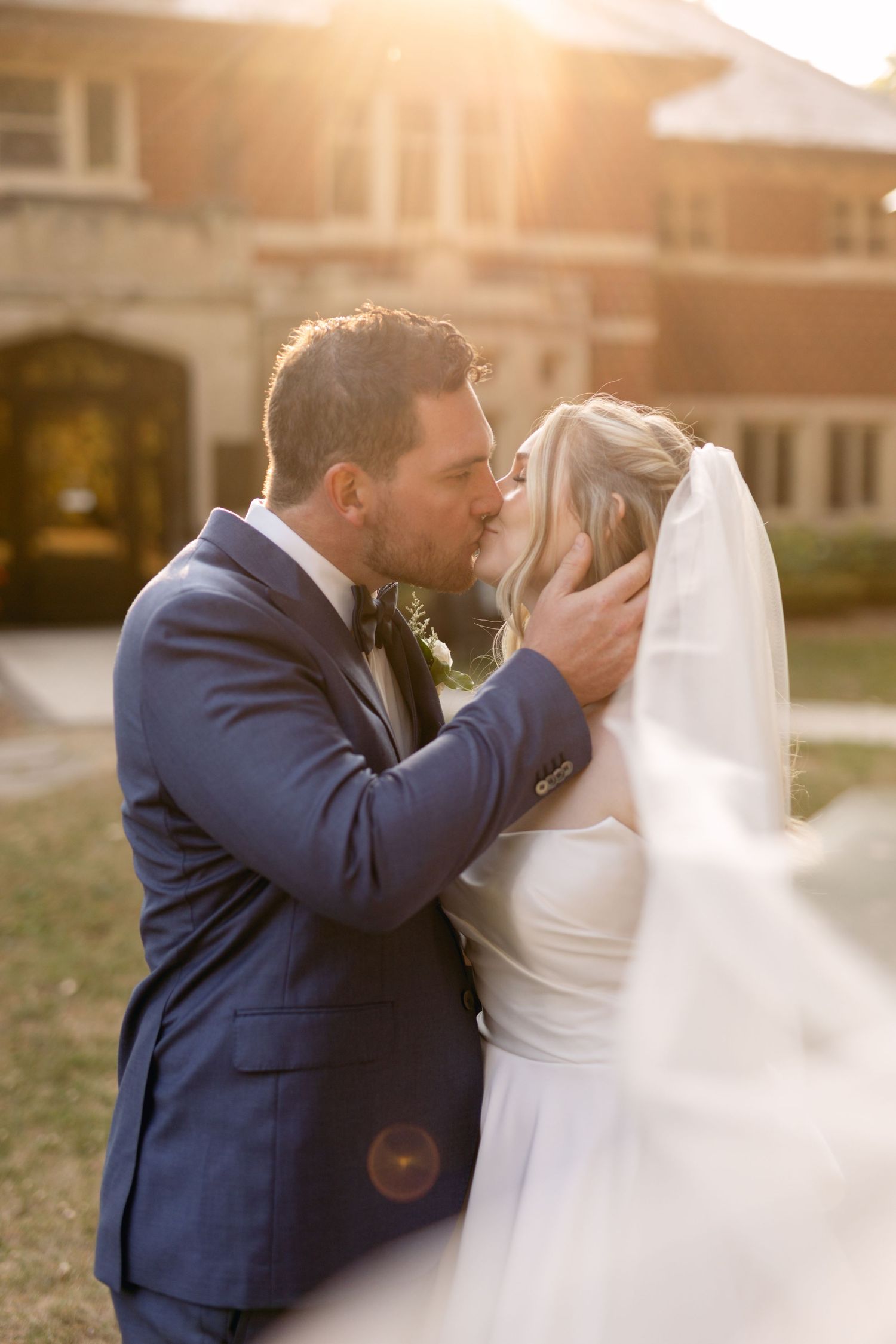 Newlyweds share an intimate moment at sunset against a brick building backdrop.