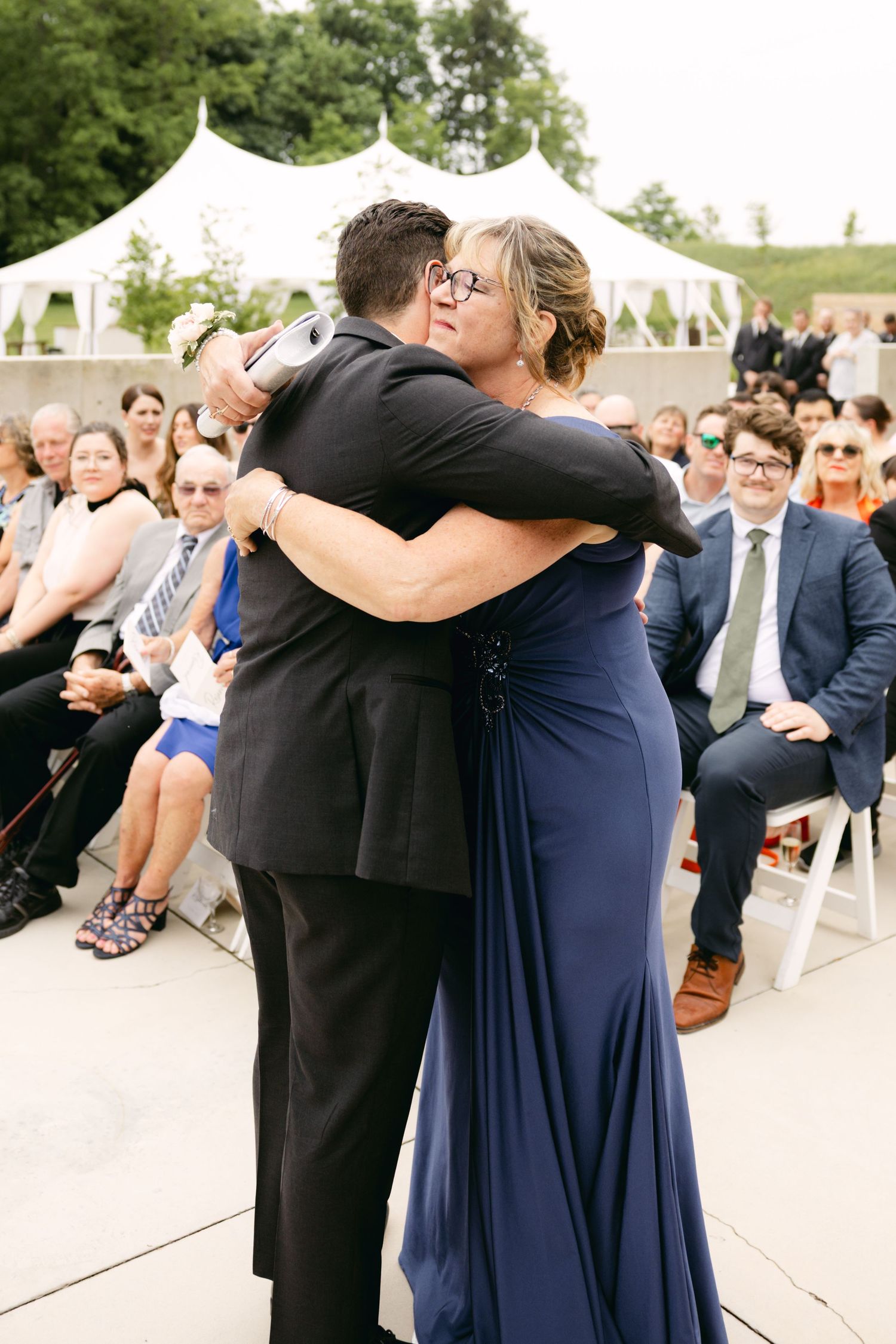 Wedding guests in formal attire embracing during outdoor celebration.