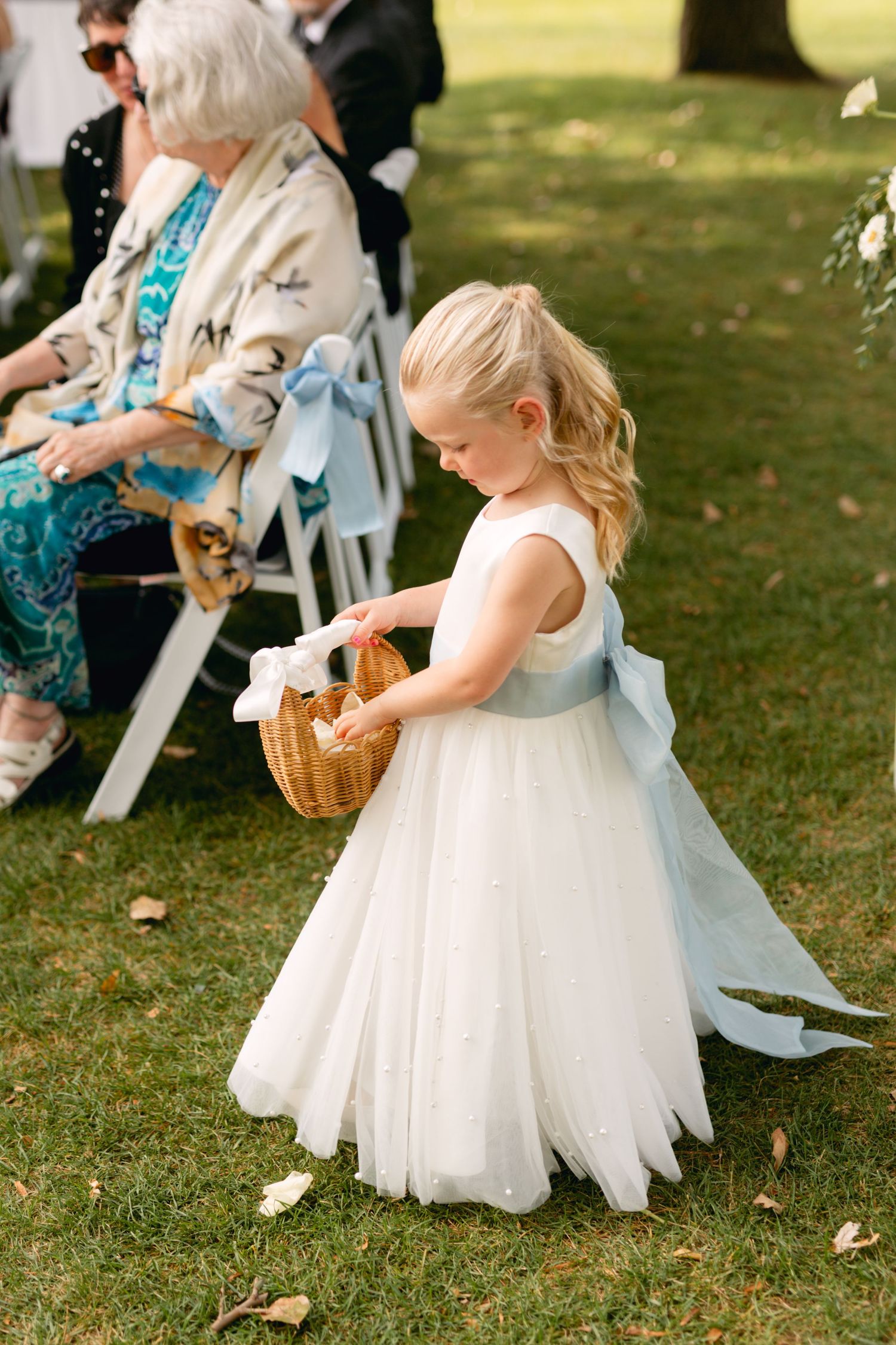 Young flower girl in white dress drops petals from basket onto grassy lawn during wedding.