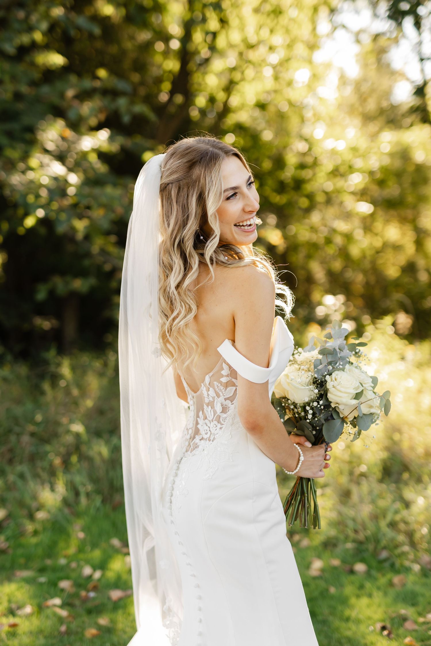 Bride holds white bouquet in elegant lace-detailed dress against garden backdrop.