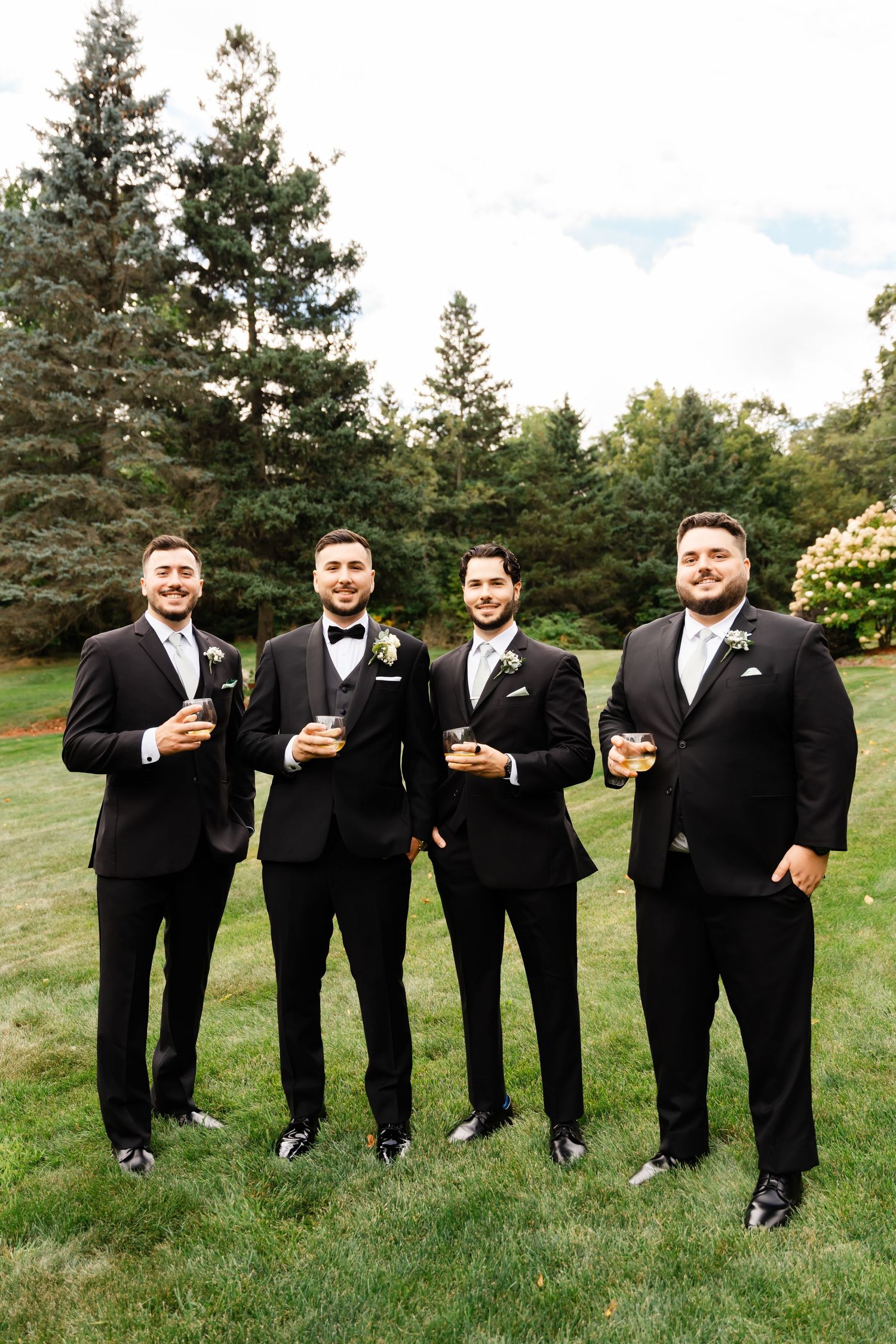 Groomsmen in black tuxedos standing together on grass with pine trees in background.