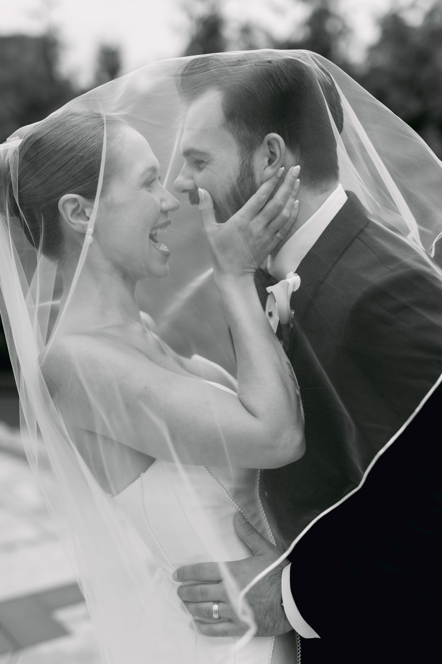 Black and white photo of bride and groom sharing intimate moment under flowing wedding veil.