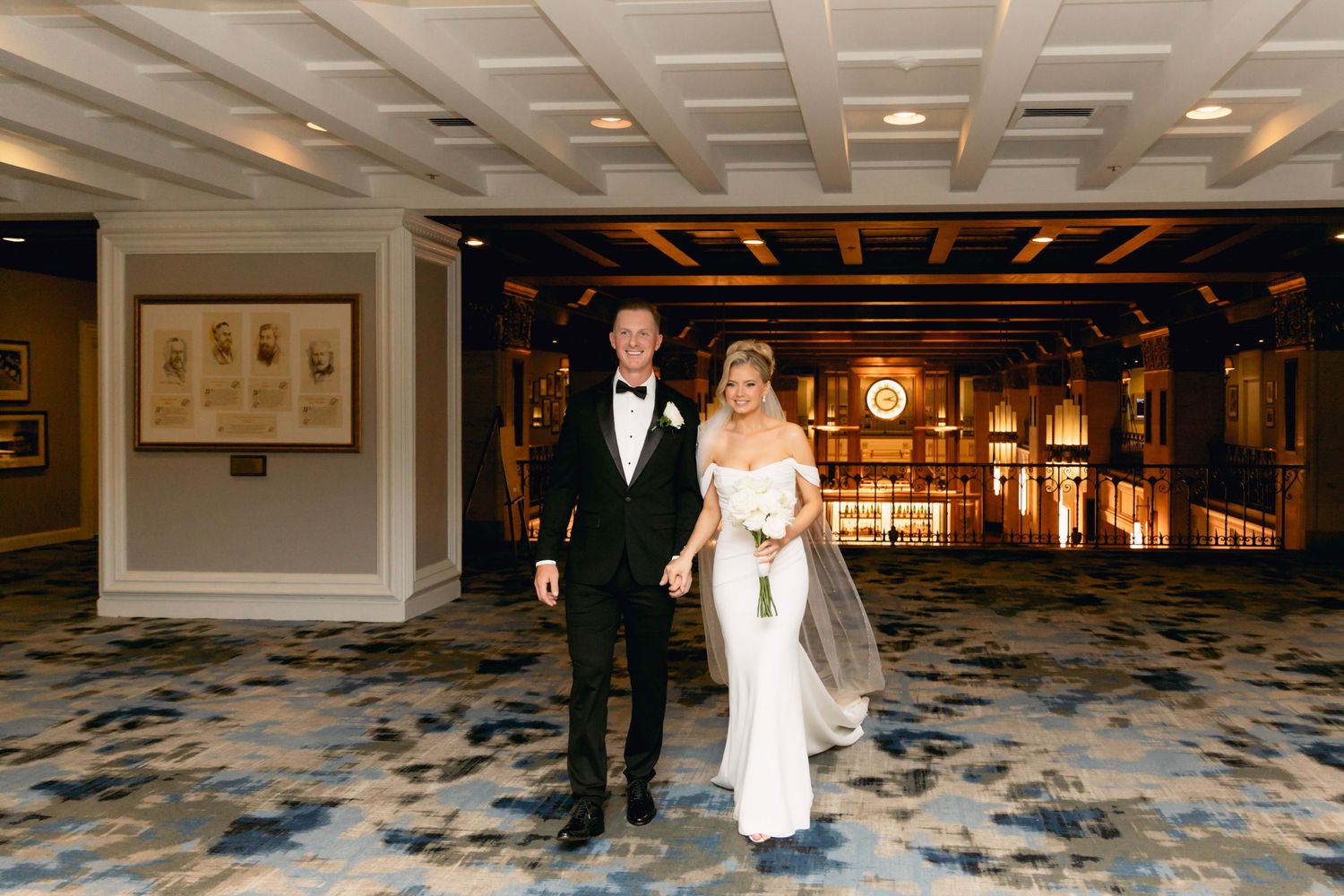 Bride and groom walk hand in hand through elegant hotel lobby with ornate decor and patterned carpet.