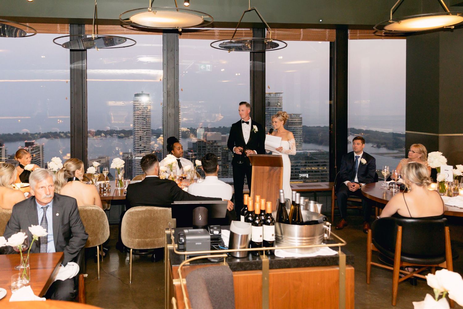 Bride and groom giving speeches at intimate rooftop wedding reception with stunning city skyline views.