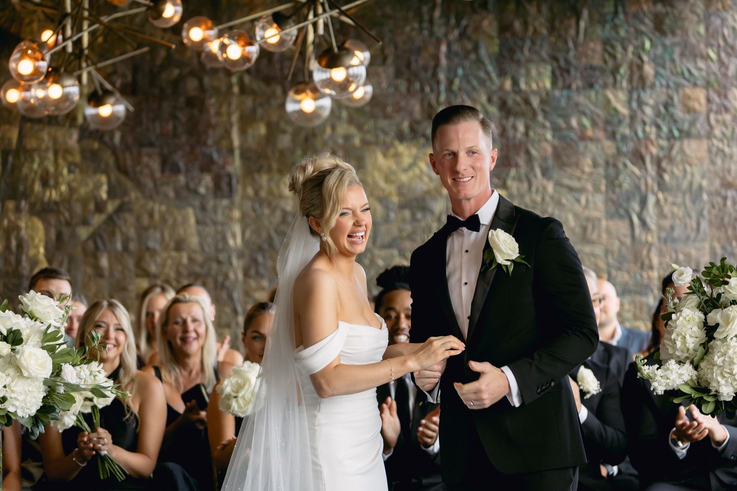 Smiling bride and groom exchange rings at altar surrounded by white floral arrangements and joyful wedding guests.
