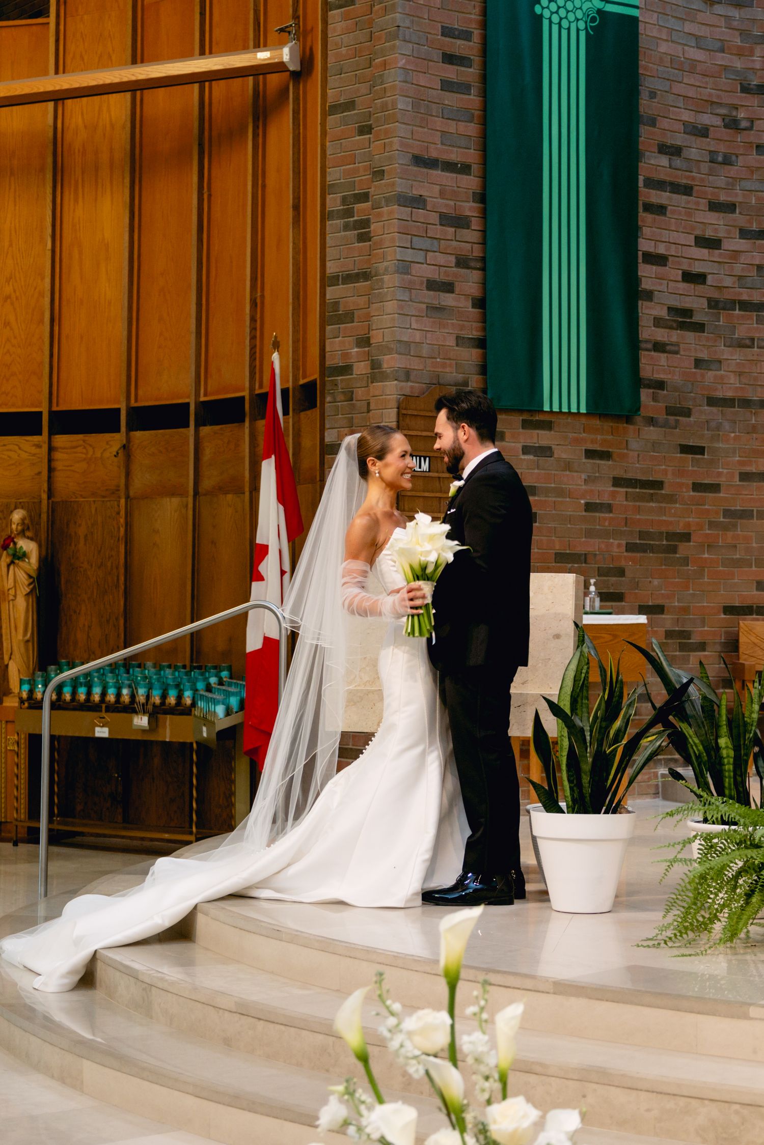 Bride in white gown with veil and groom in black tuxedo facing each other at church altar.
