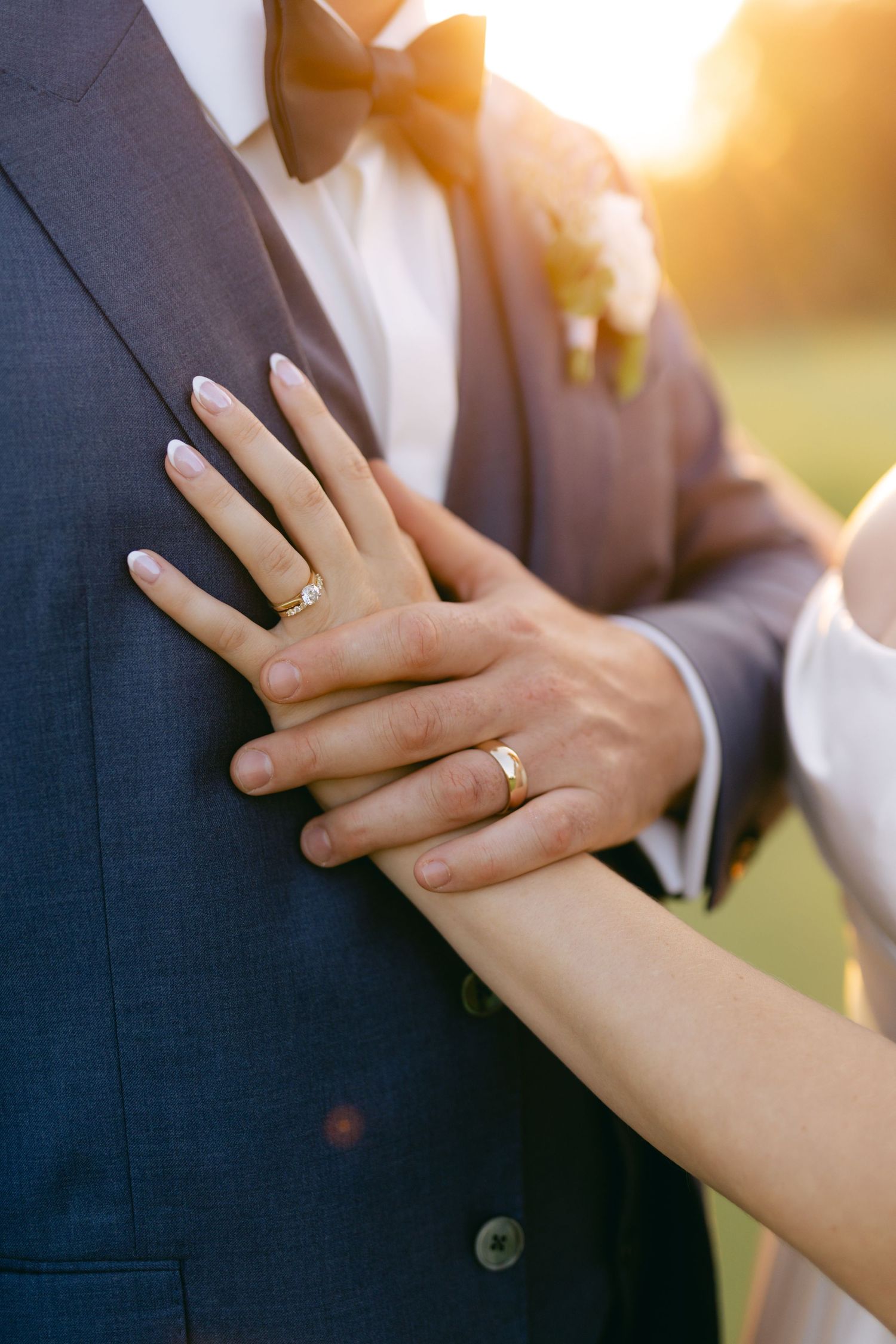 Close-up of wedding rings and hands against formal attire in warm sunset light.