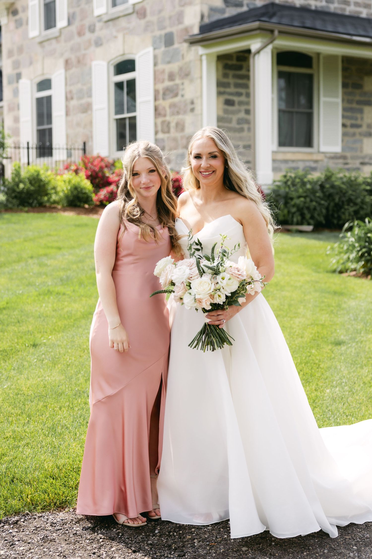 Bride in white gown and bridesmaid in pink dress hold bouquets while standing on grass near stone building.