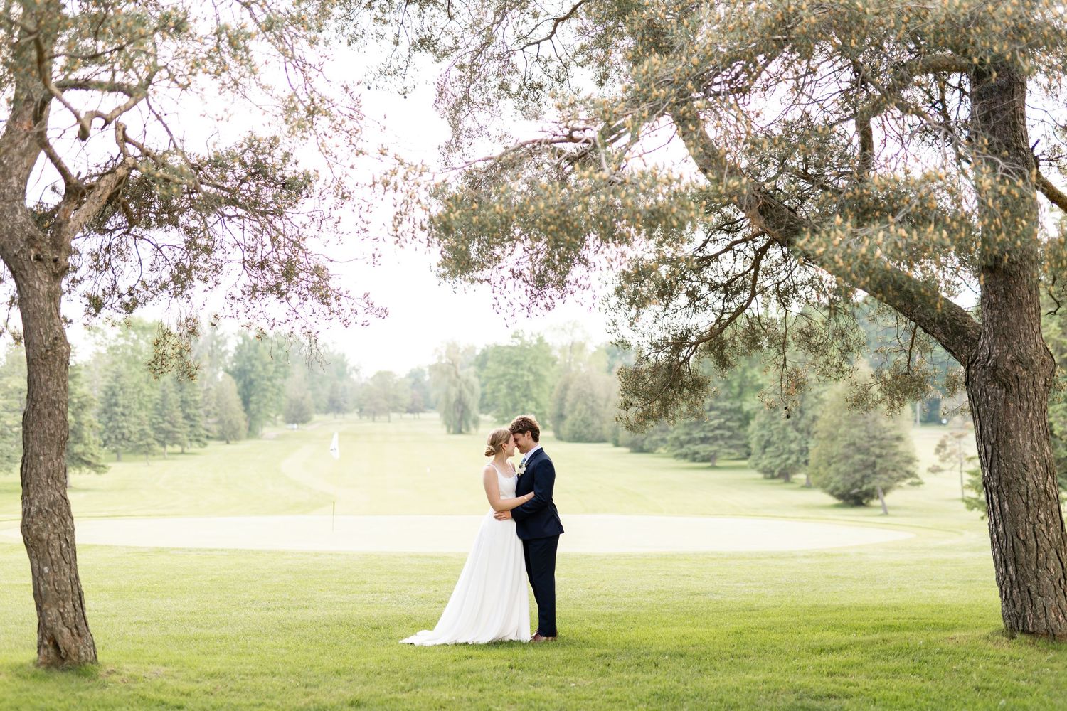 Newlyweds embrace on a rolling green lawn with trees in the background.