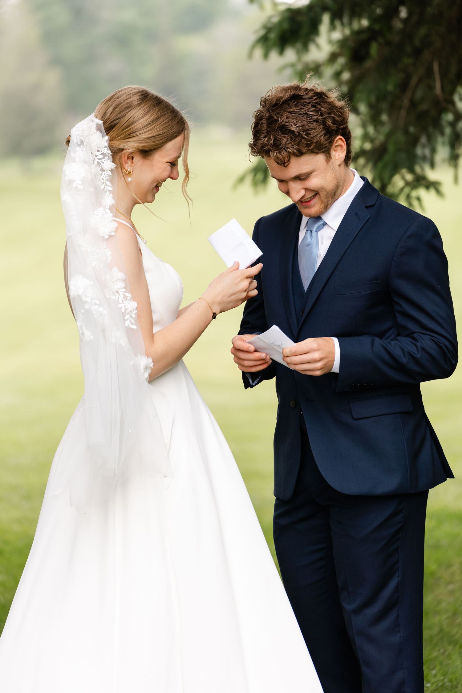 A bride in a white lace dress and groom in navy suit share a private moment reading notes in an outdoor garden setting.