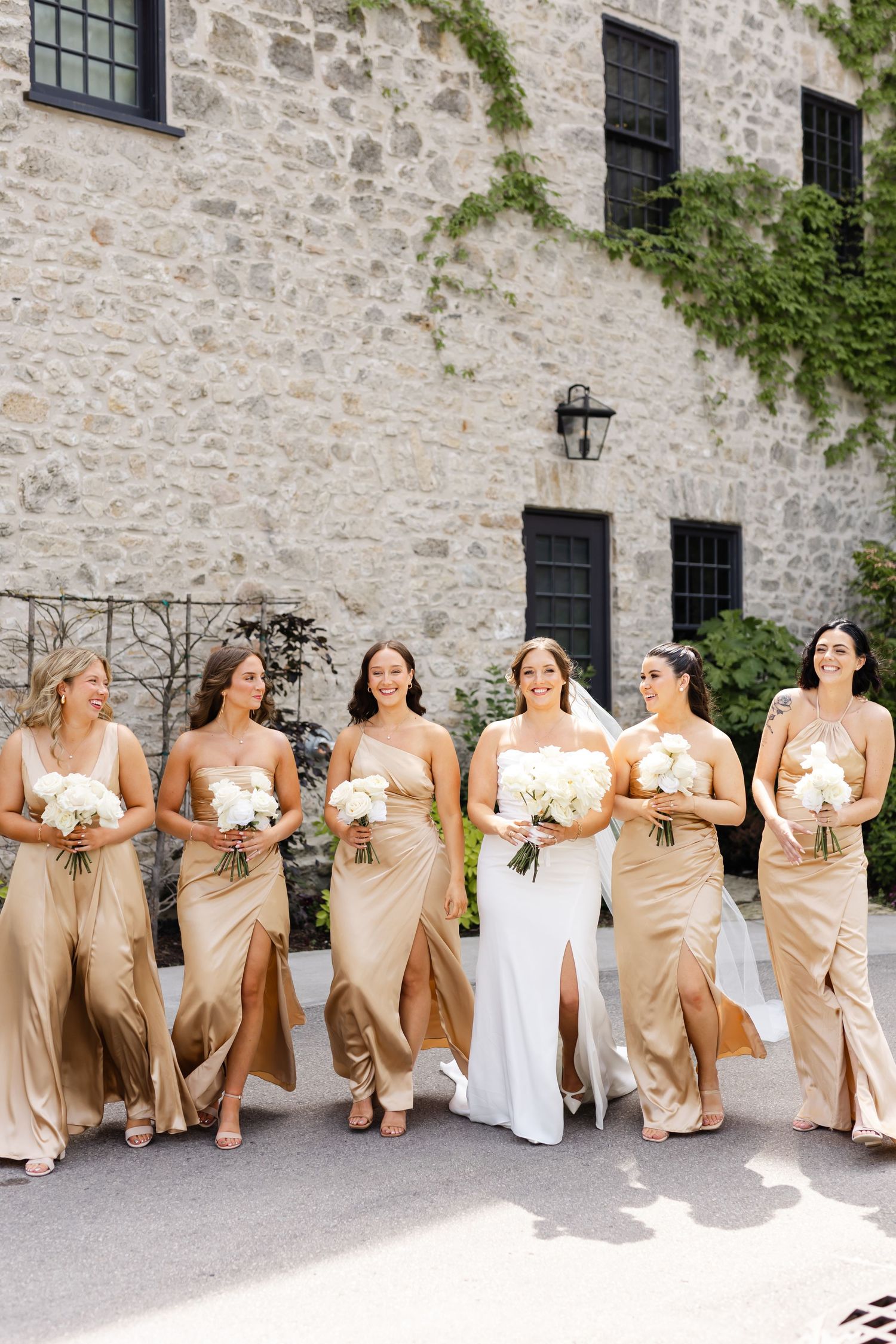 Bridal party in gold satin gowns and bride in white dress walk together holding white floral bouquets outdoors.