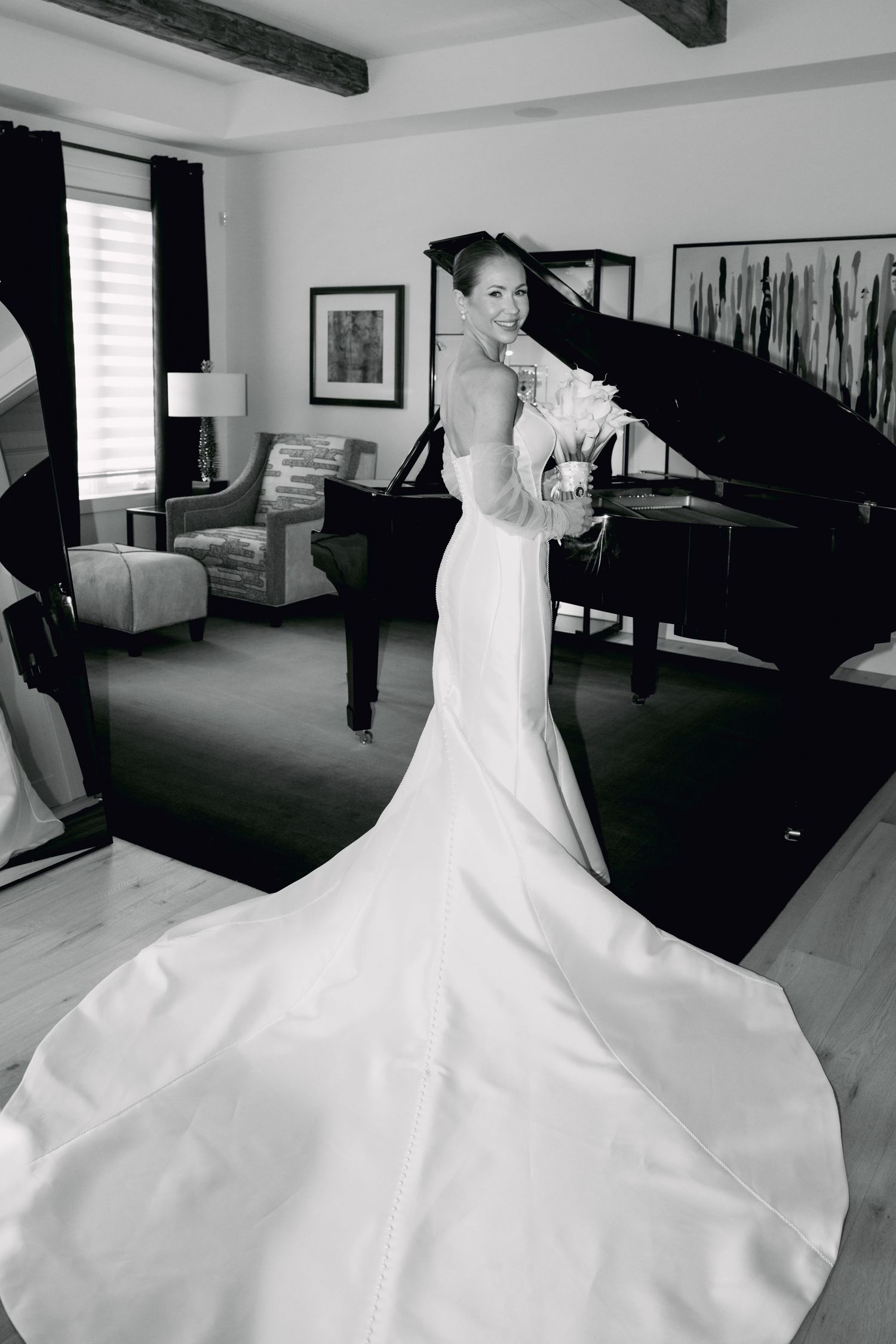 Elegant bride in fitted white gown with long train poses by grand piano in black and white photo.