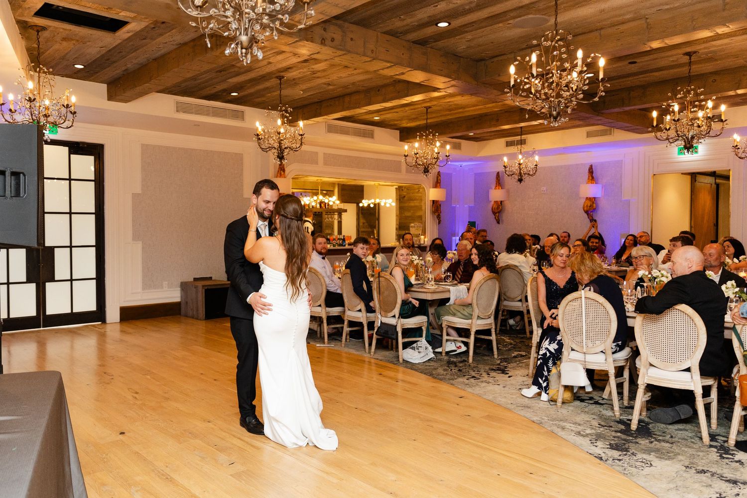 Bride and groom share first dance in elegant reception hall with chandeliers and seated guests.