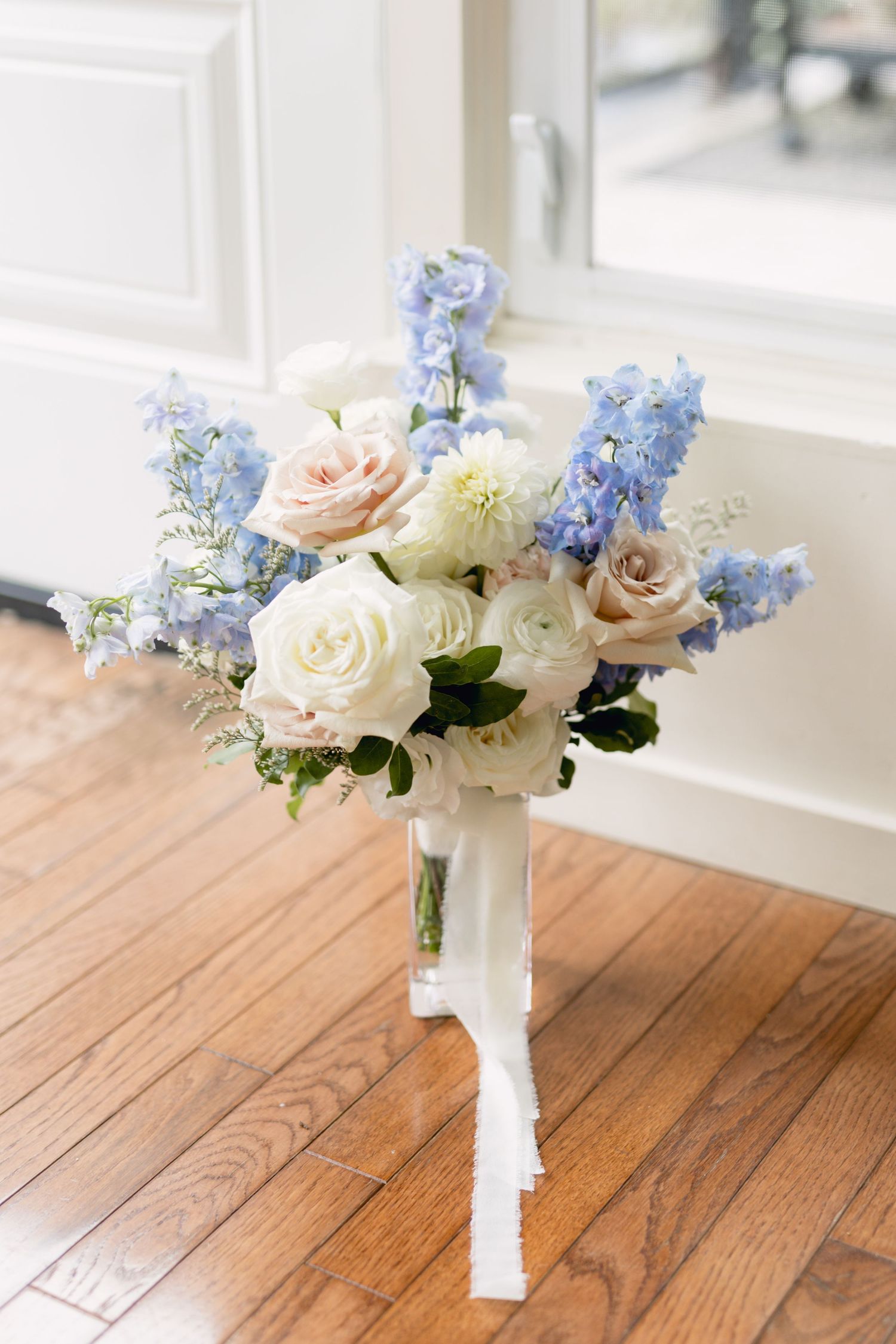 Elegant floral arrangement with roses and blue delphiniums in white vase on wooden table near window.