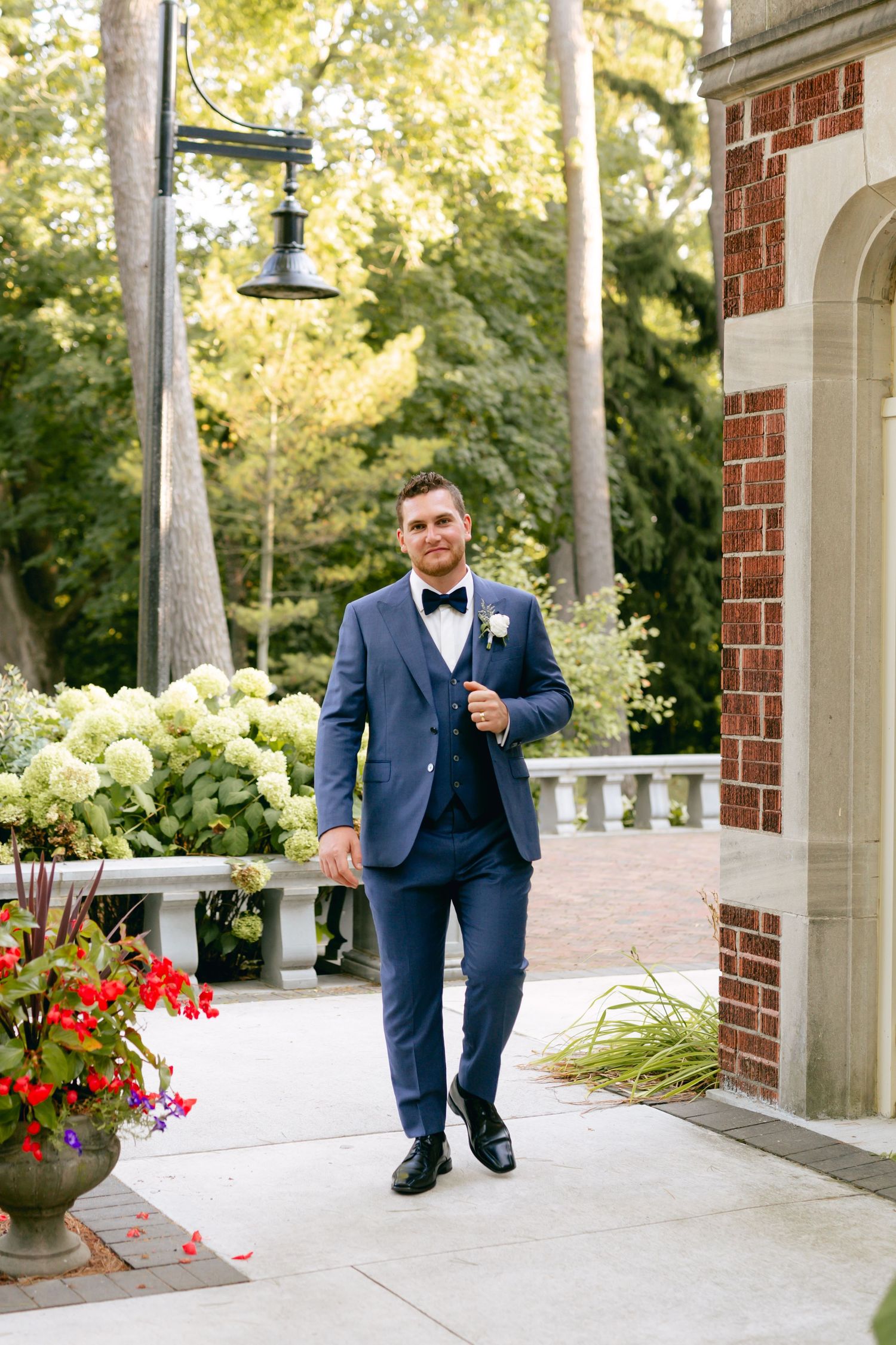 Groom in navy suit walks along a columned walkway with red flowers and greenery.