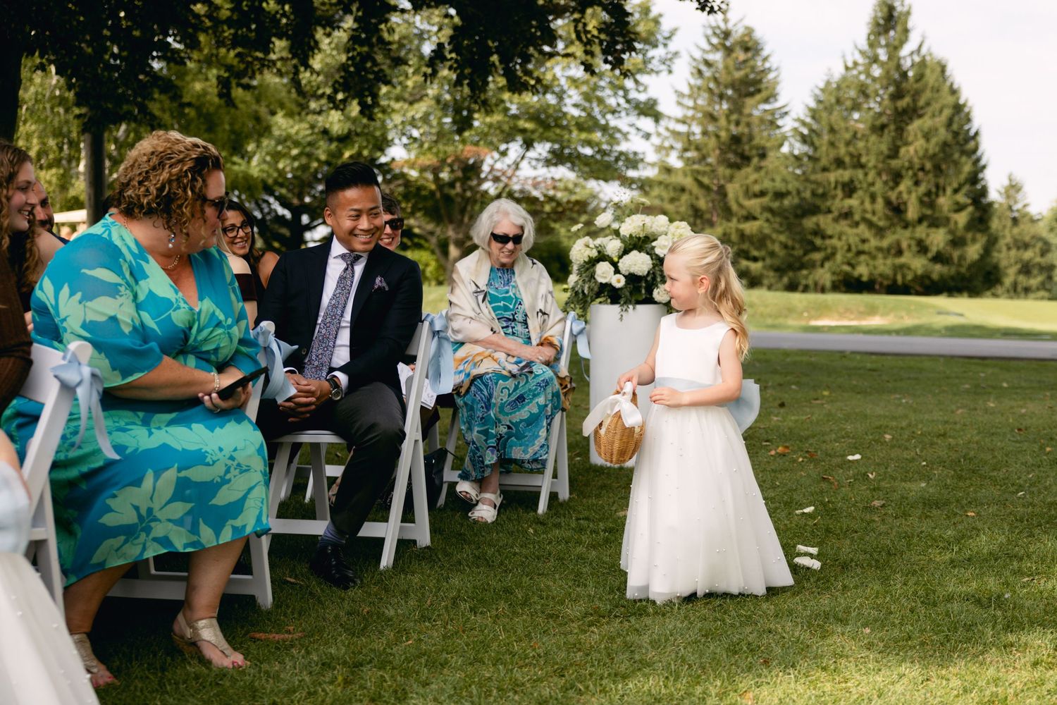 A flower girl in a white dress scatters petals during an outdoor wedding ceremony.
