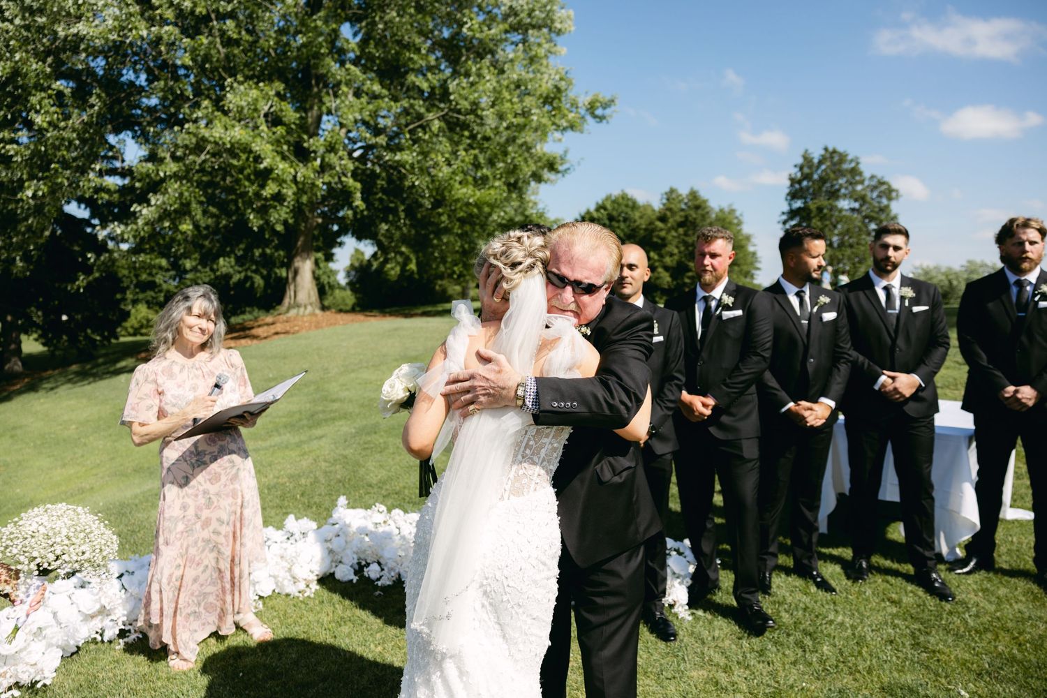 Wedding party and guests gather for group photos on a sunny lawn with trees in background.