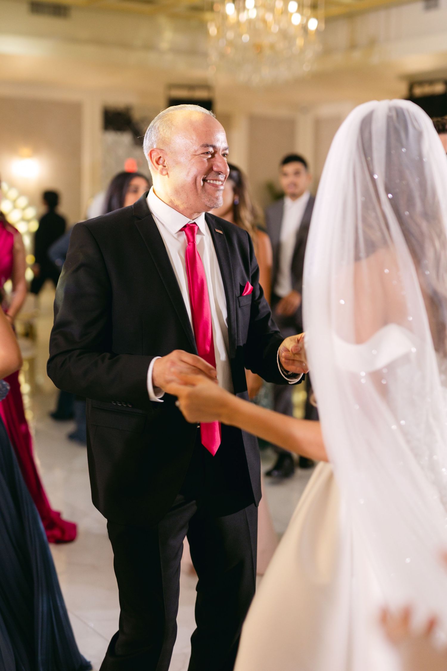 A guest in a black suit and red tie dances with the bride at an elegant wedding reception.