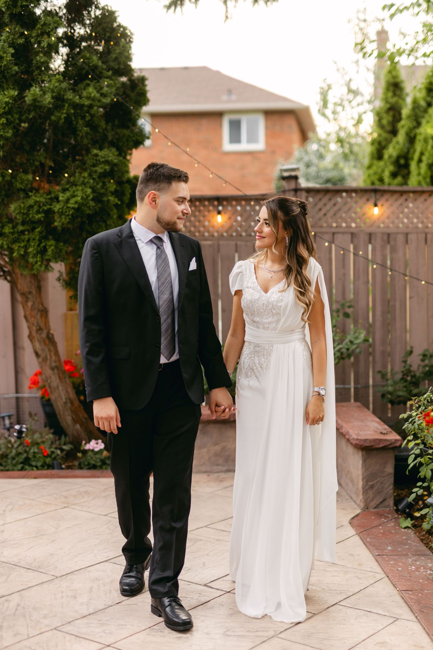 Wedding couple walking together on patio with string lights and wooden fence in background.