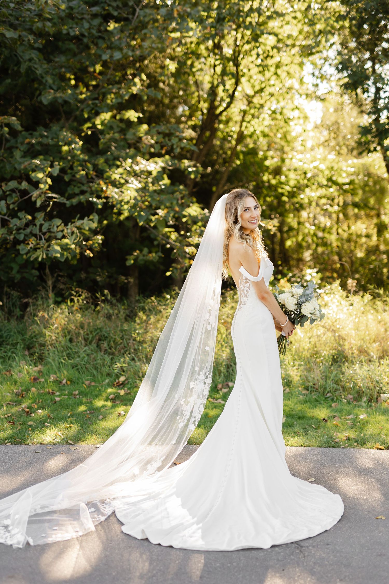 Bride in fitted white gown with cathedral veil poses on outdoor path.